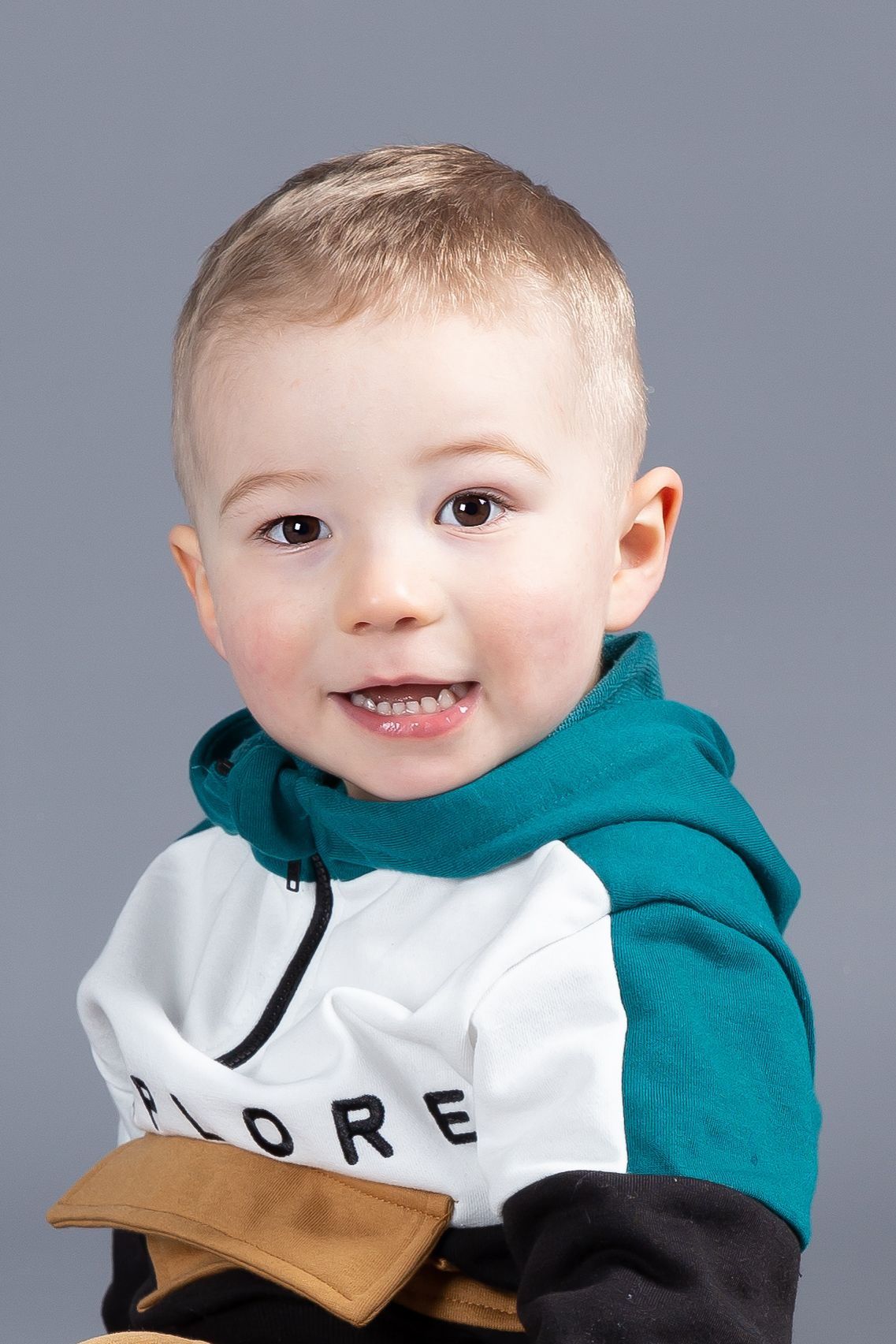 Young child smiles while wearing a white and teal hooded sweatshirt with text against a gray background.