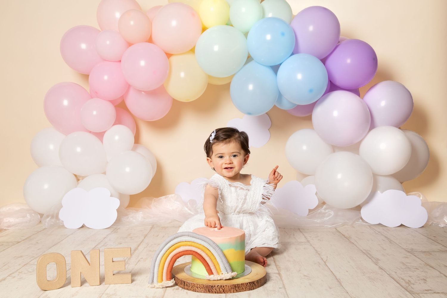 Rainbow pastel balloons and decorations create a whimsical first birthday photo session setup on light wooden floor.
