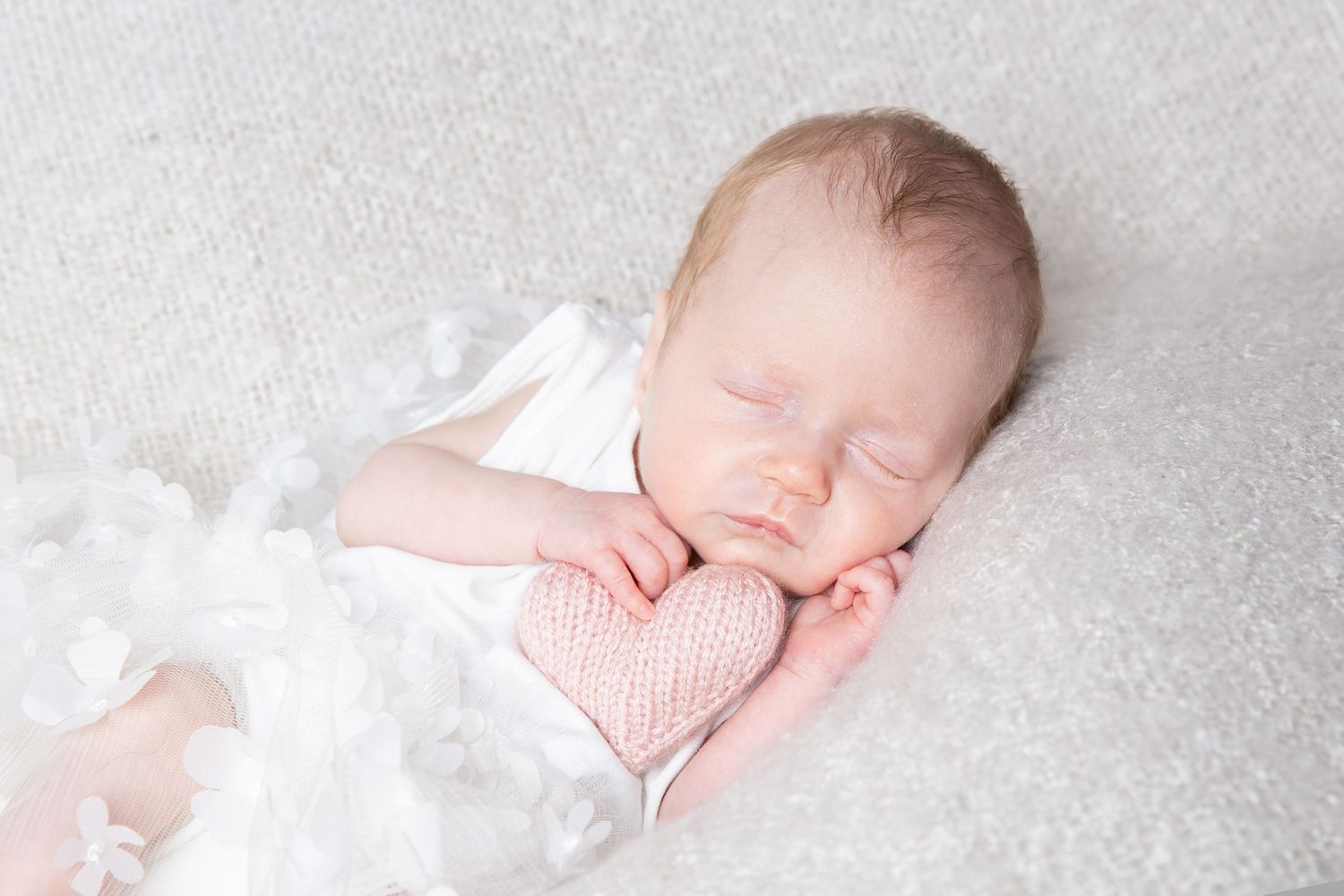 Peaceful sleeping newborn baby wearing pink wrapped in white blanket on soft white backdrop.