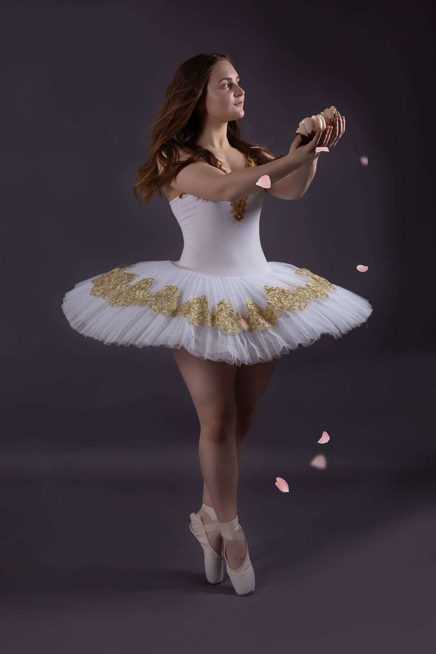 Ballet dancer in white classical tutu with gold accents performs graceful poses on dark backdrop with scattered pink petals.