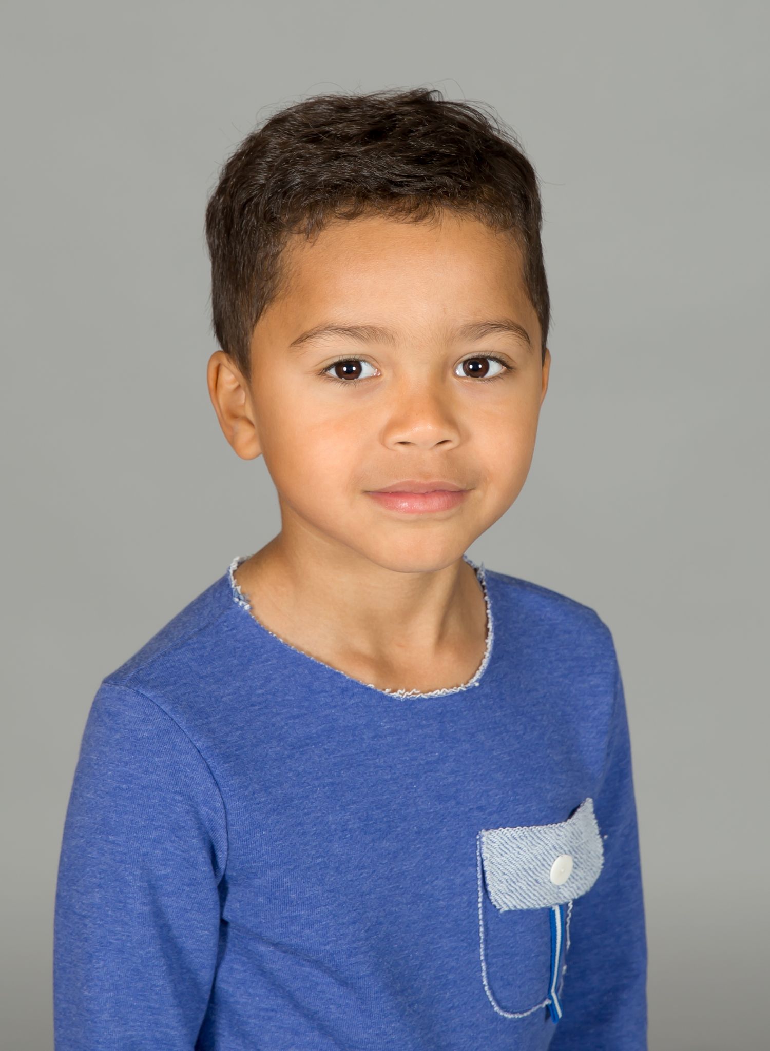 Young child wearing a blue t-shirt with a small pocket detail smiles at the camera against a gray background.