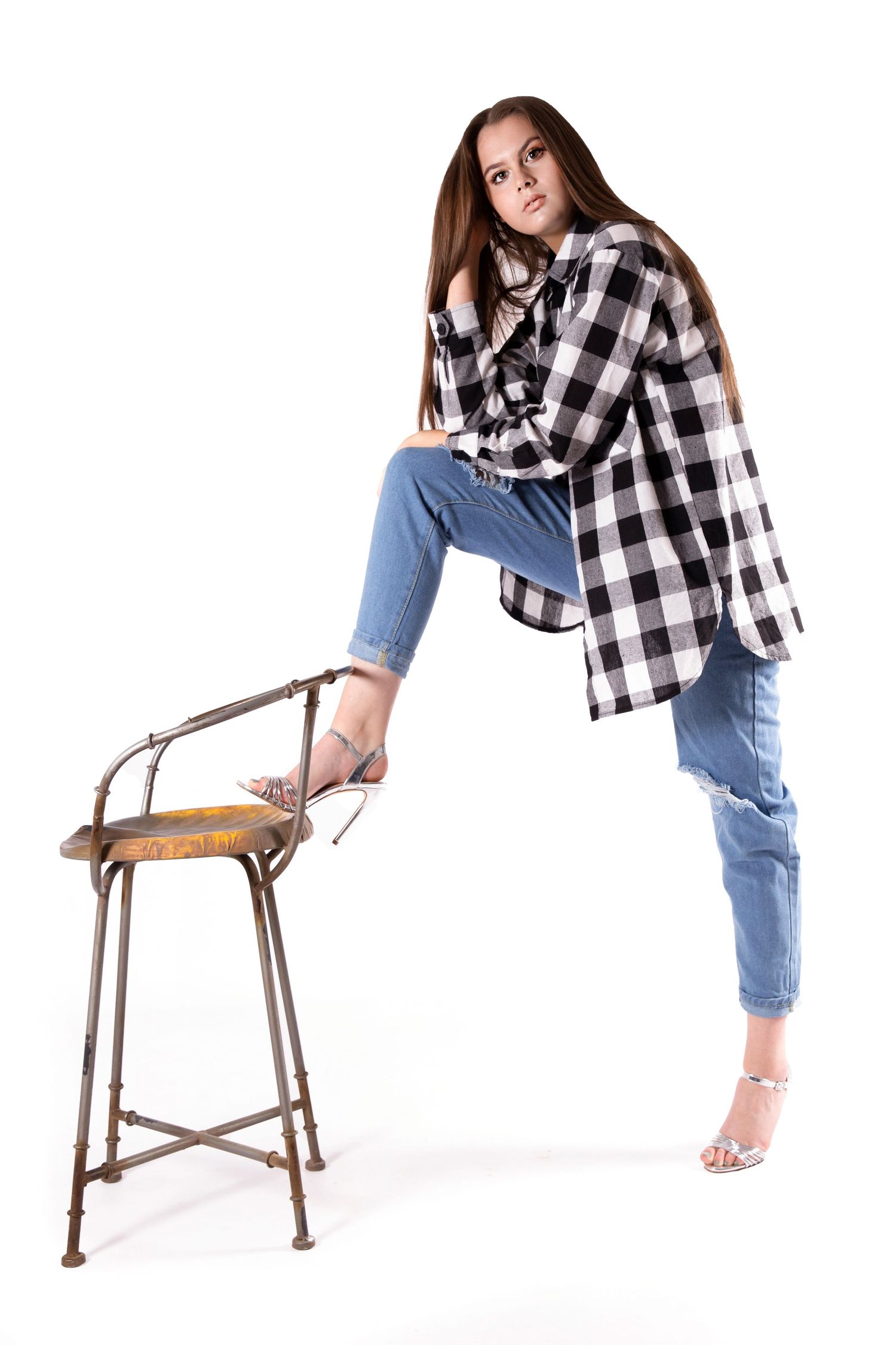 Model in black and white checkered shirt and cropped jeans poses with vintage metal stool against white background.