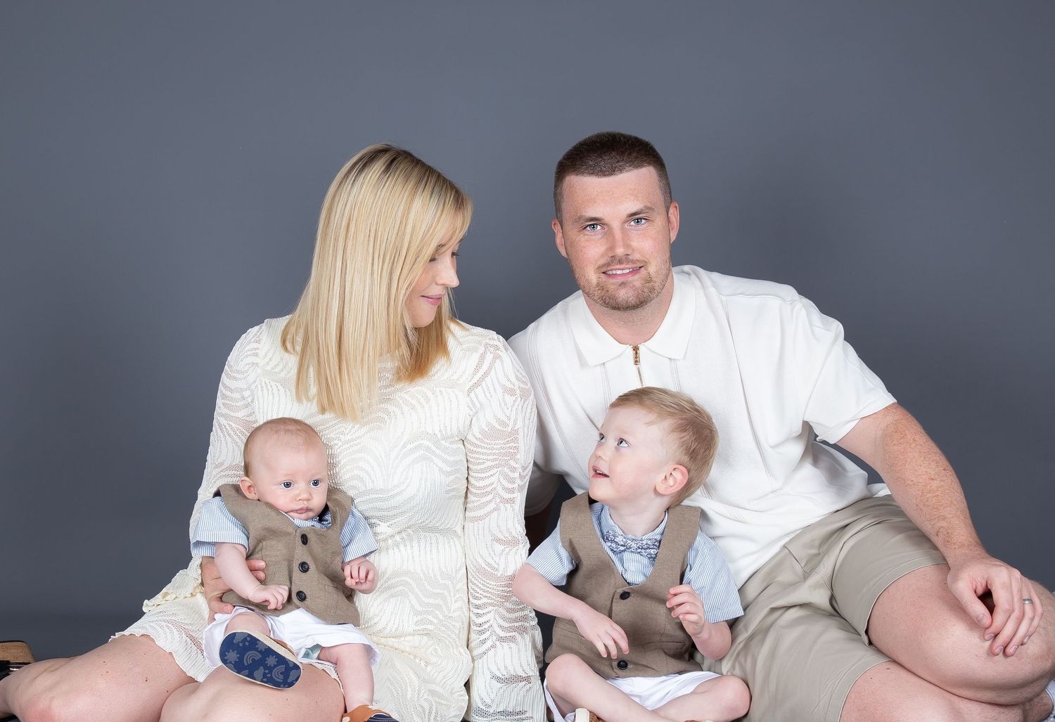 Family wearing coordinated white outfits poses together for a series of casual studio portraits against a grey backdrop.