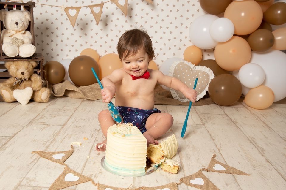 A cake smash photo session setup with brown and white balloons and a small white cake on a polka dot backdrop.