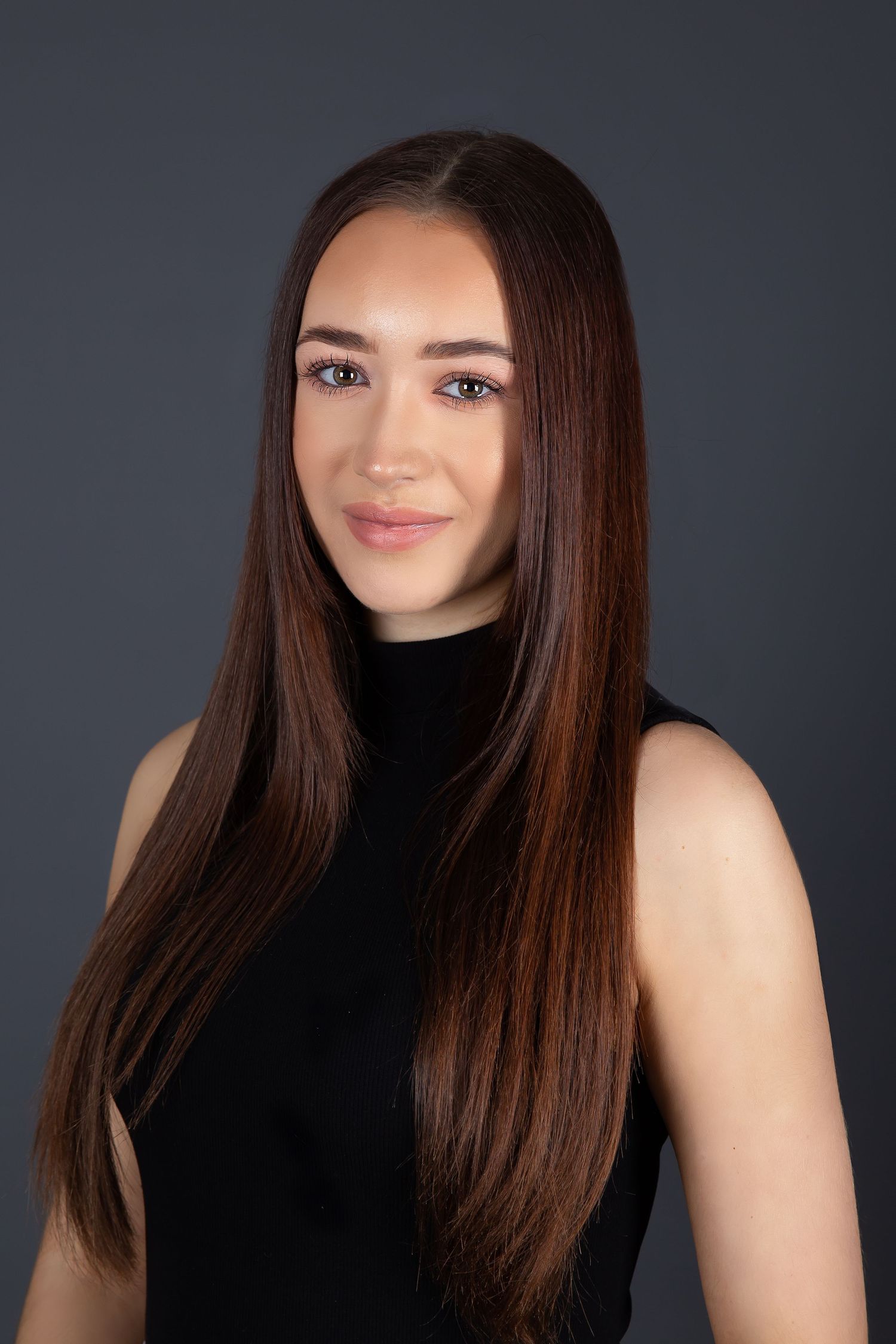 Portrait of someone with long straight dark brown hair wearing a black sleeveless top against a dark gray background.