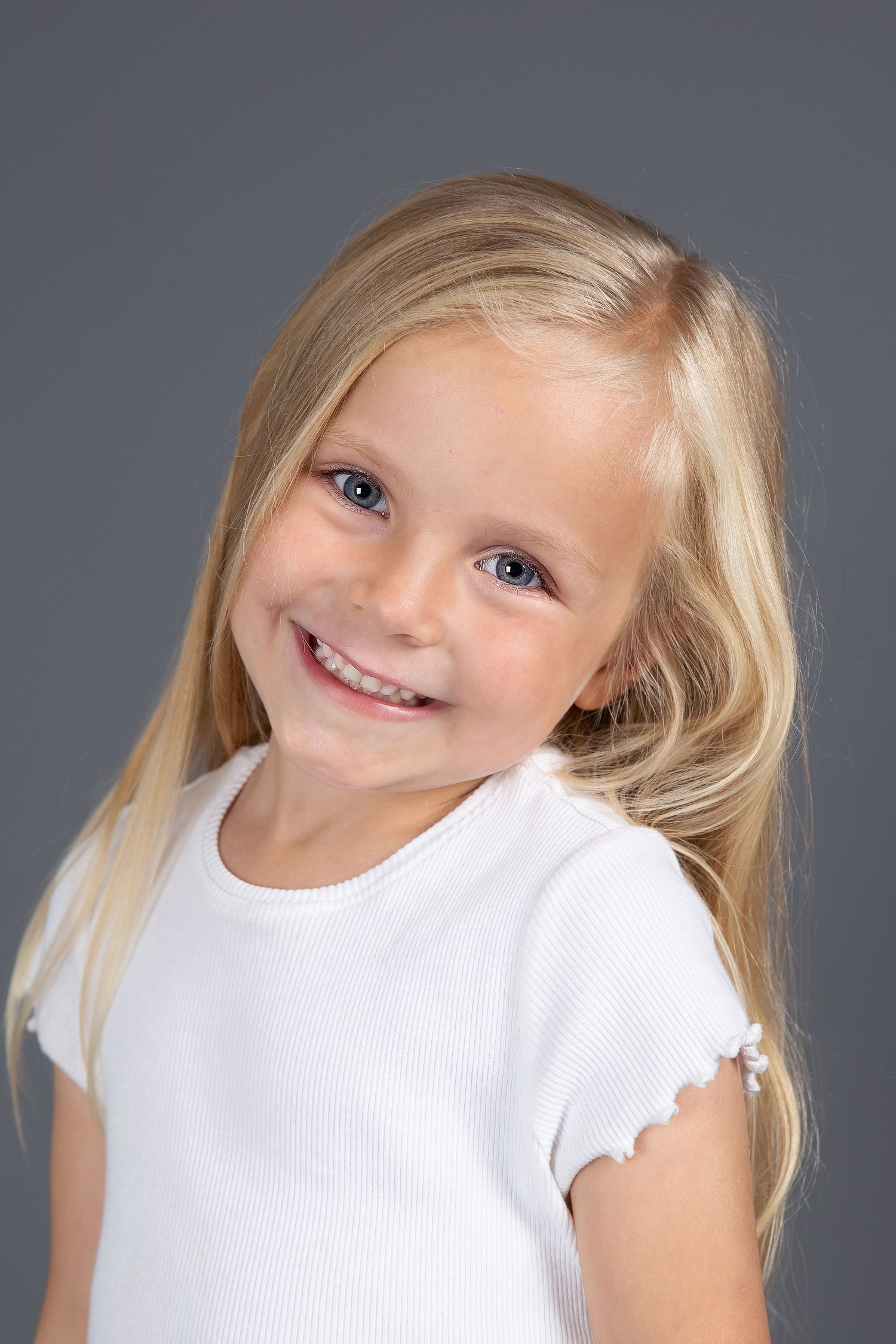 A young blonde child in a white shirt smiles brightly against a gray background.