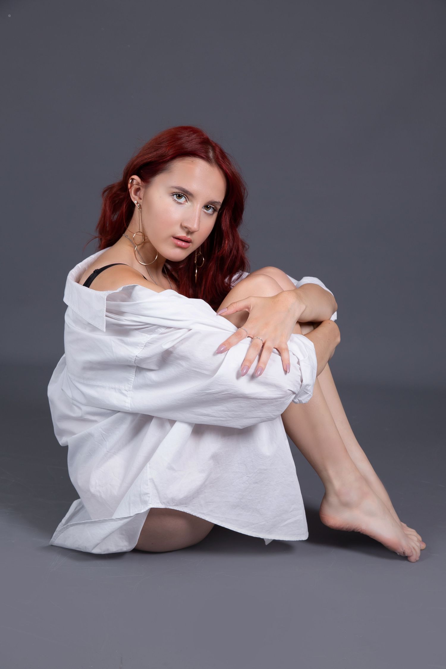 Woman with red hair in white shirt sits on floor against gray background.