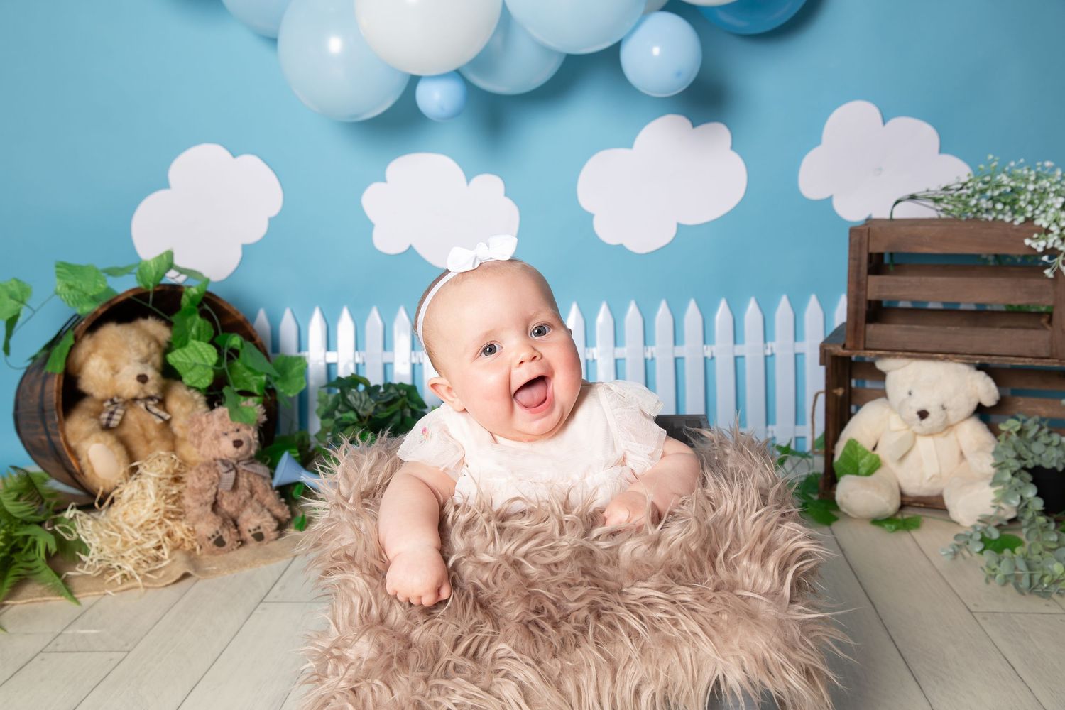 Baby wearing a soft floral headband lies on brown fur with white picket fence and cloud decorations in the background.