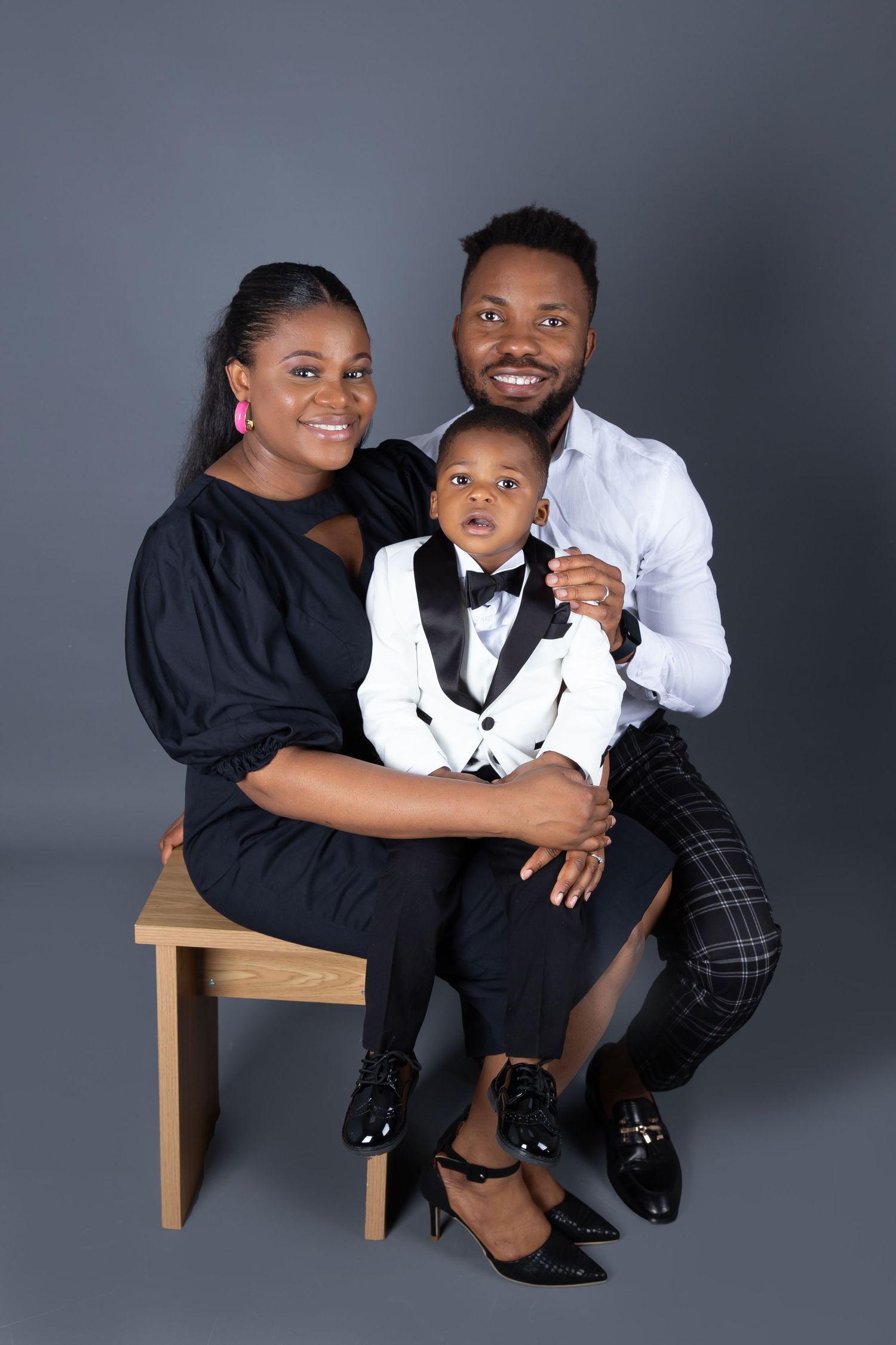 Beautiful family portrait with parents and toddler son in formal attire posing on wooden bench against gray backdrop.