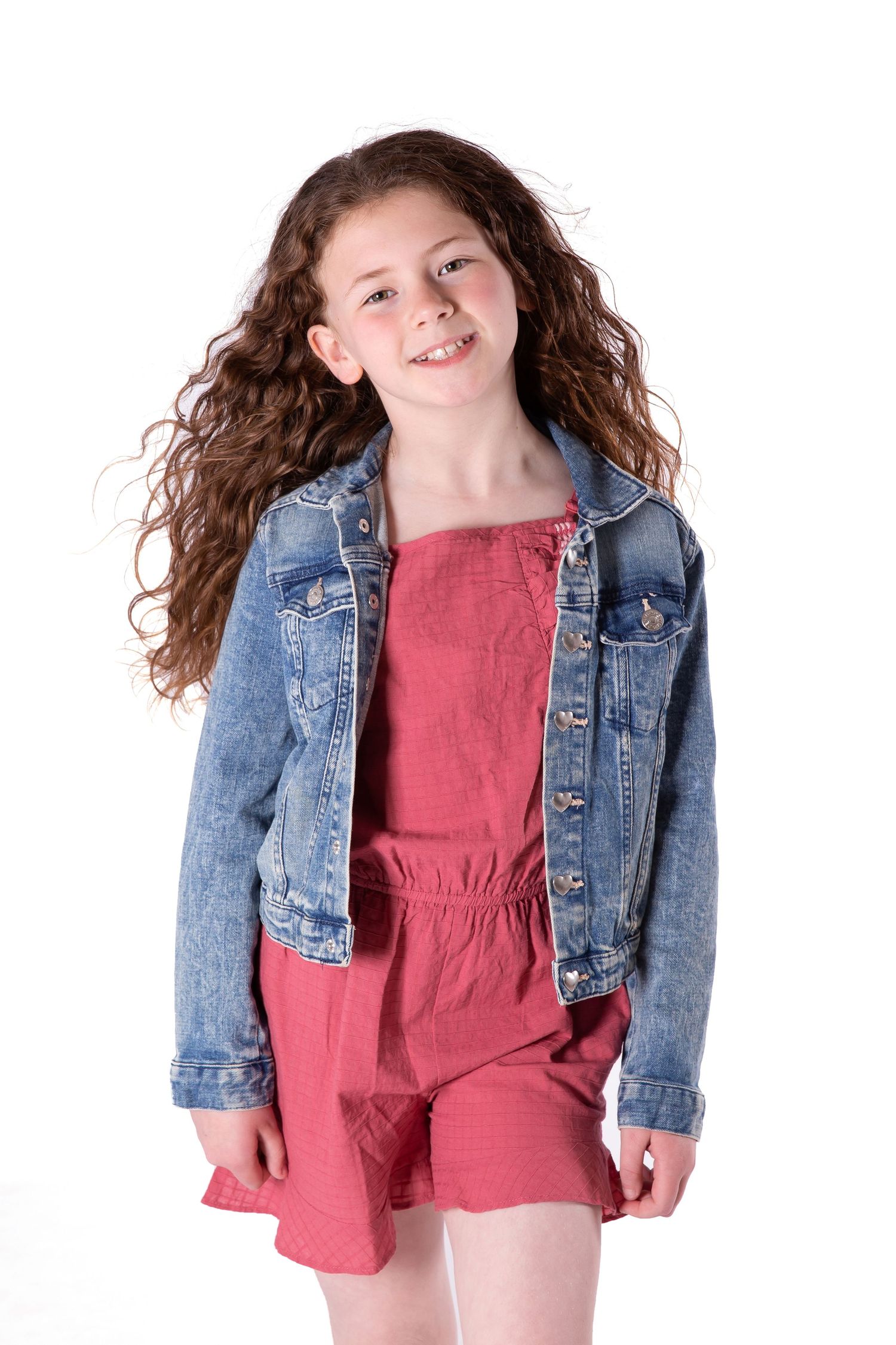 Joyful model wearing a pink romper and denim jacket twirls with curly hair against white background.