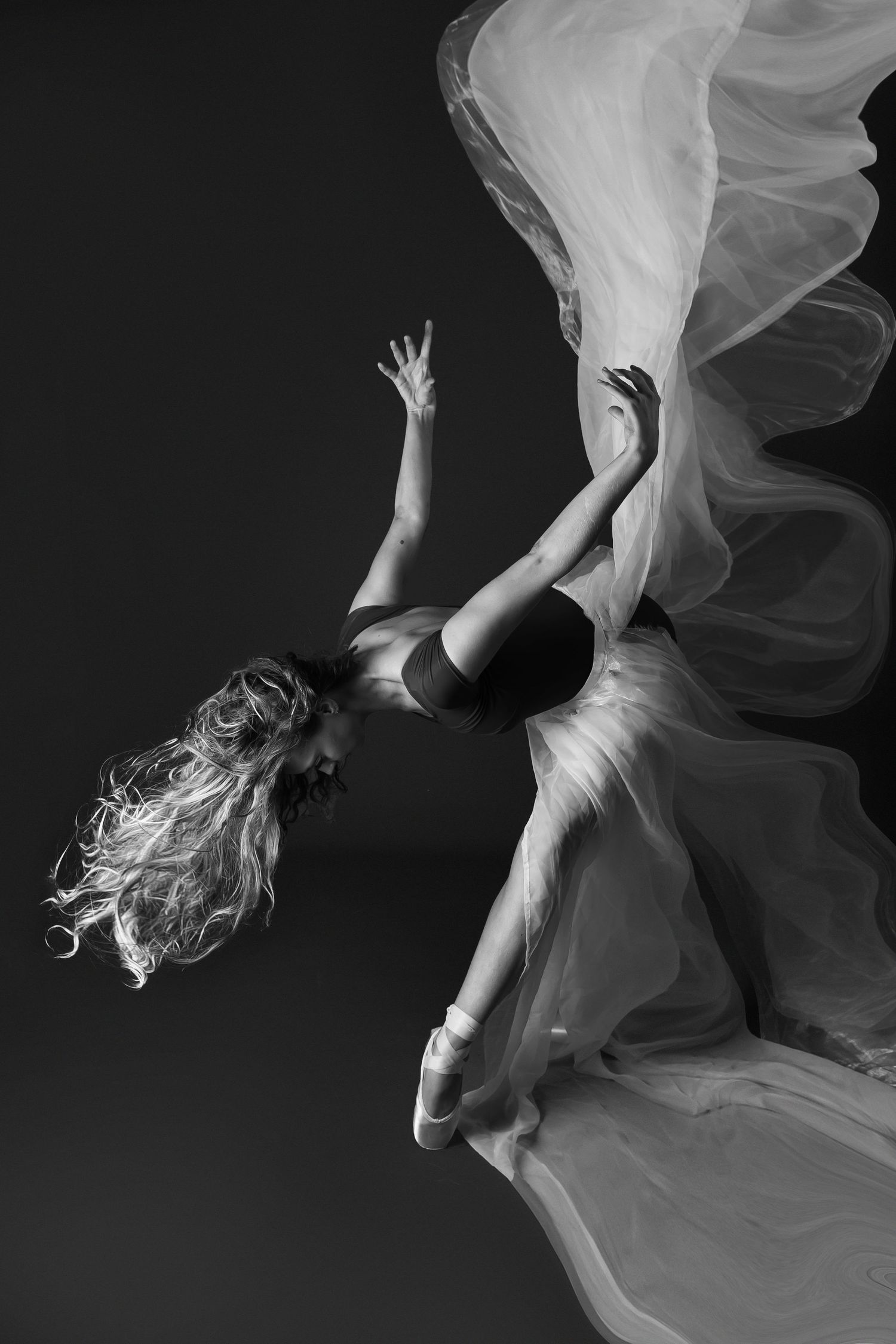 Dramatic black and white photo of a ballet dancer leaping with flowing white fabric and long hair in motion.