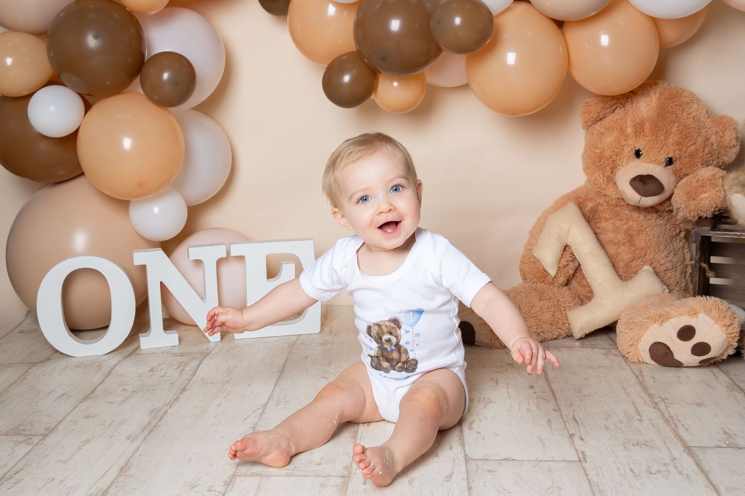 First birthday celebration setup with balloon arch, white ONE letters, and teddy bear on wooden floor.