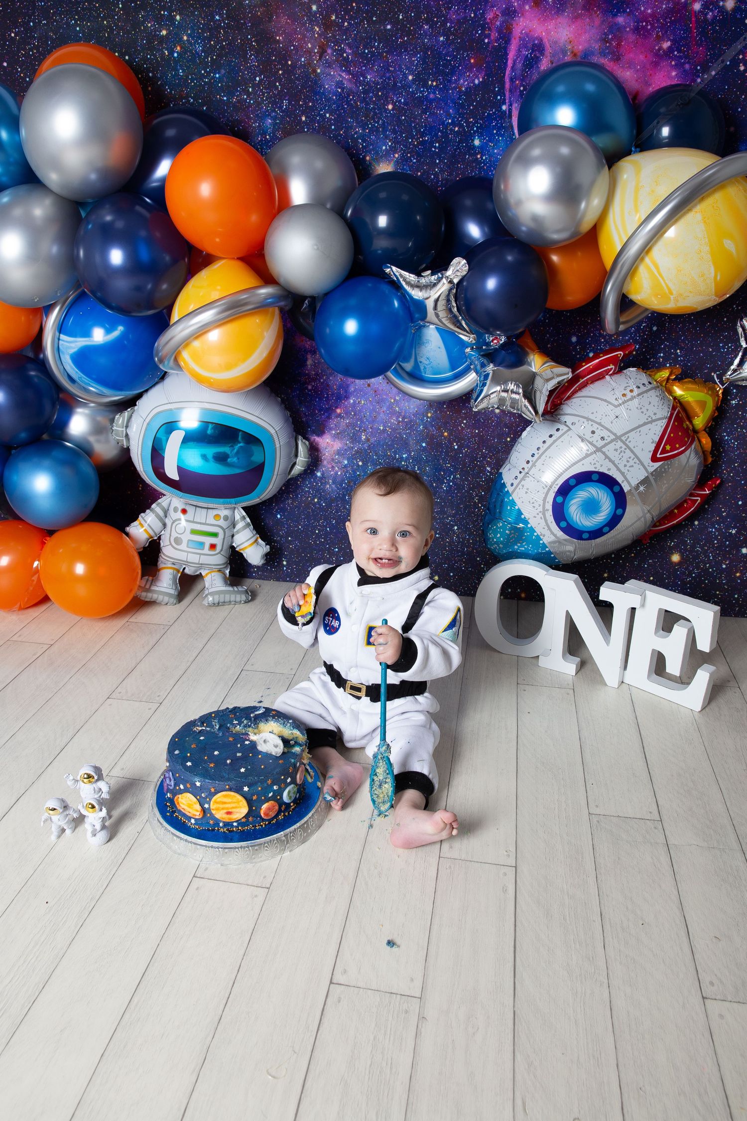 Space themed birthday party backdrop with balloons, rockets and astronaut decorations against a starry galaxy background.