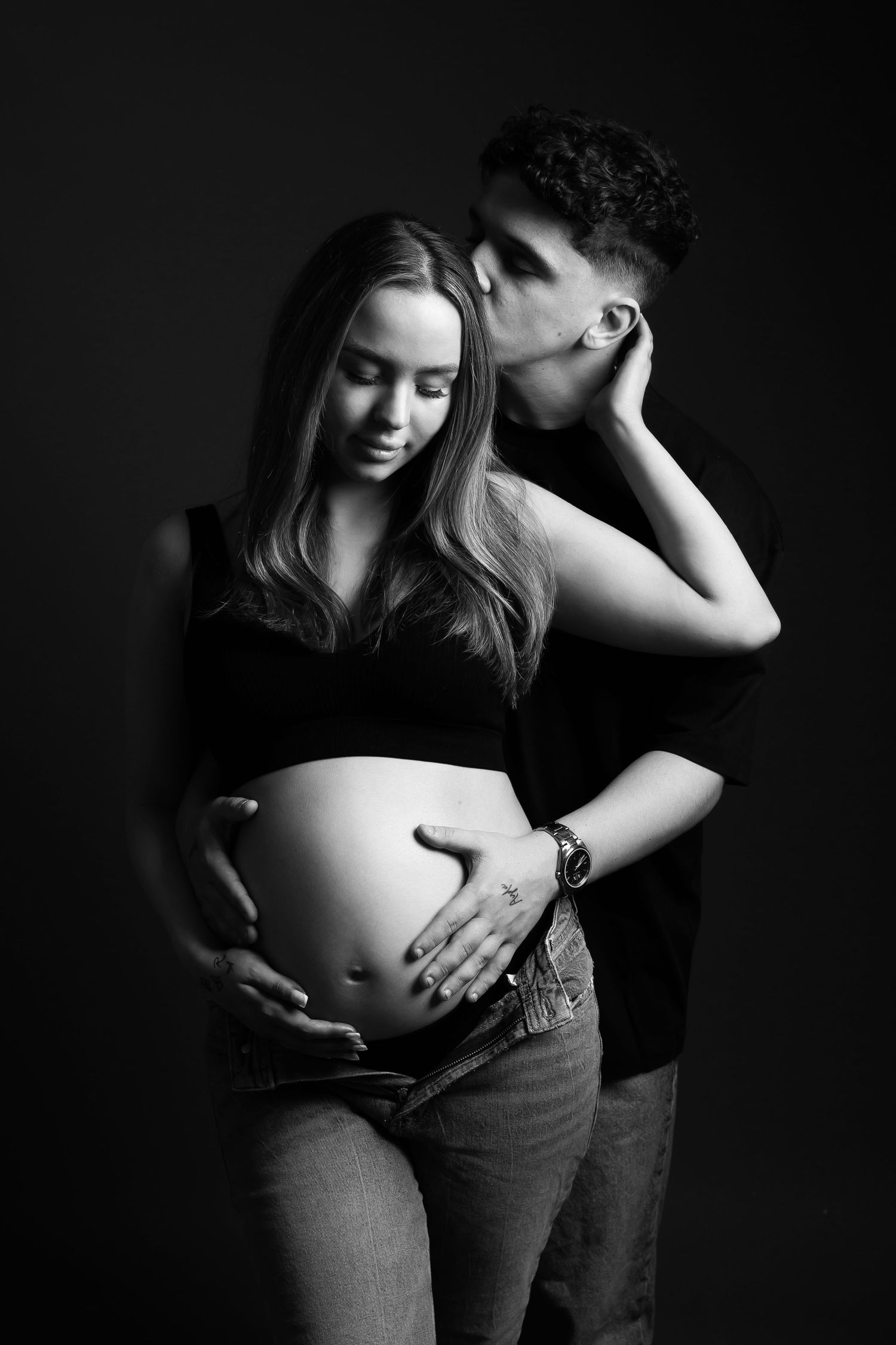 Black and white artistic maternity photo showing intimate moment between couple as they embrace expectant mother's belly.