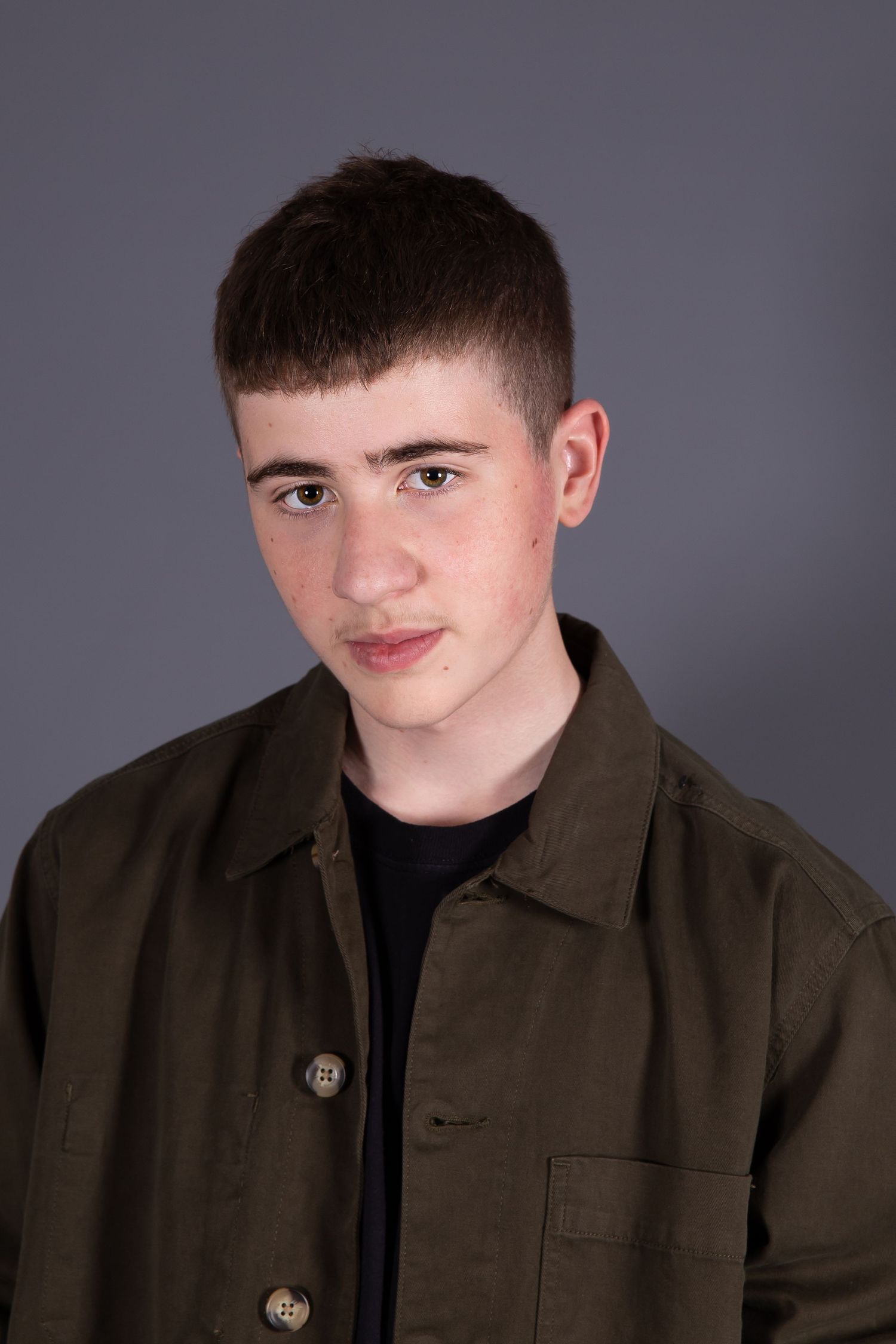 Studio portrait of a young person wearing an olive green jacket over a black shirt against a gray background.