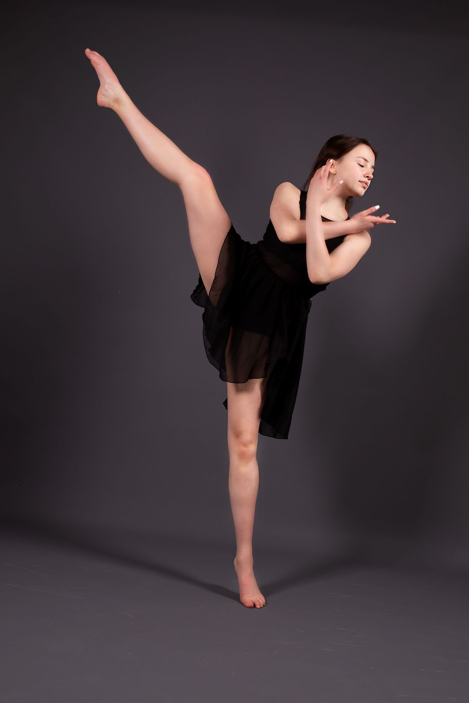 Dancer in black dress performs graceful high kick against dark background during contemporary dance pose.