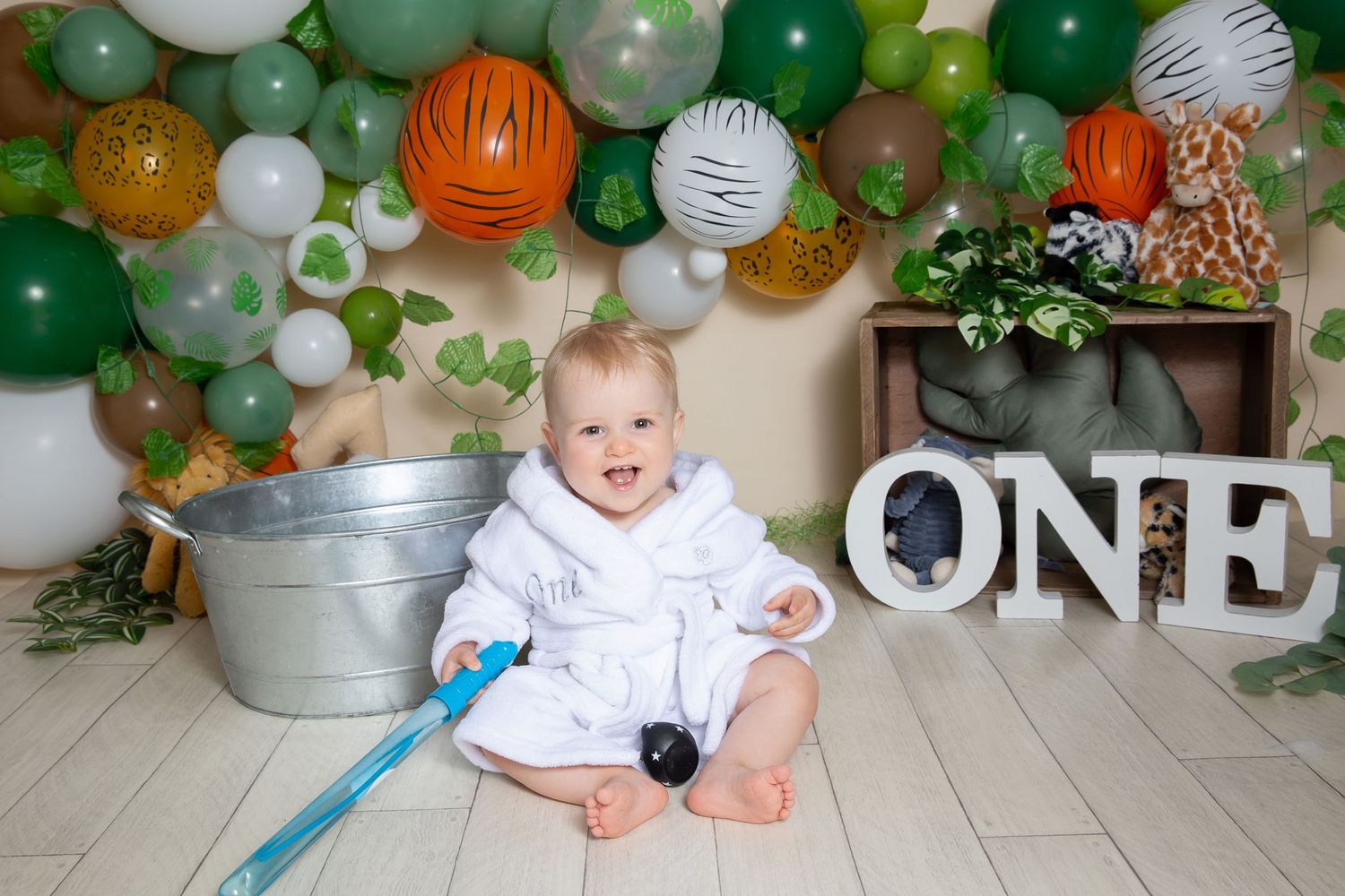 A smiling baby in white sits on a floor with green and orange balloon decorations and a 'ONE' sign for their first birthday.