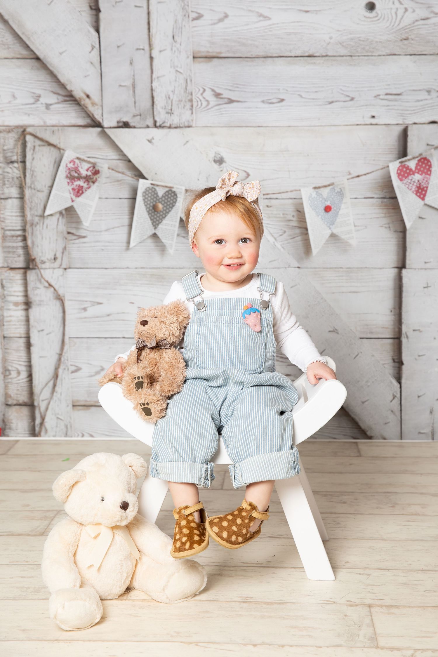 Toddler in blue overalls sits on white chair with teddy bears against rustic wall with heart banner decorations.