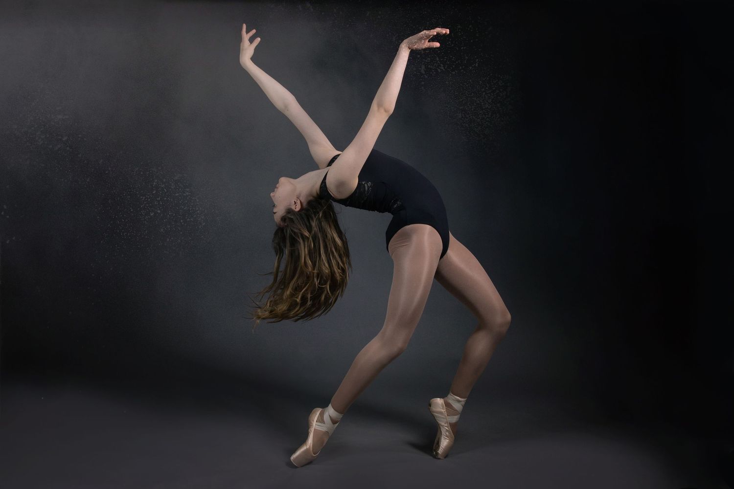 Contemporary ballet dancer in black leotard performs graceful backbend position on pointe against dark background.