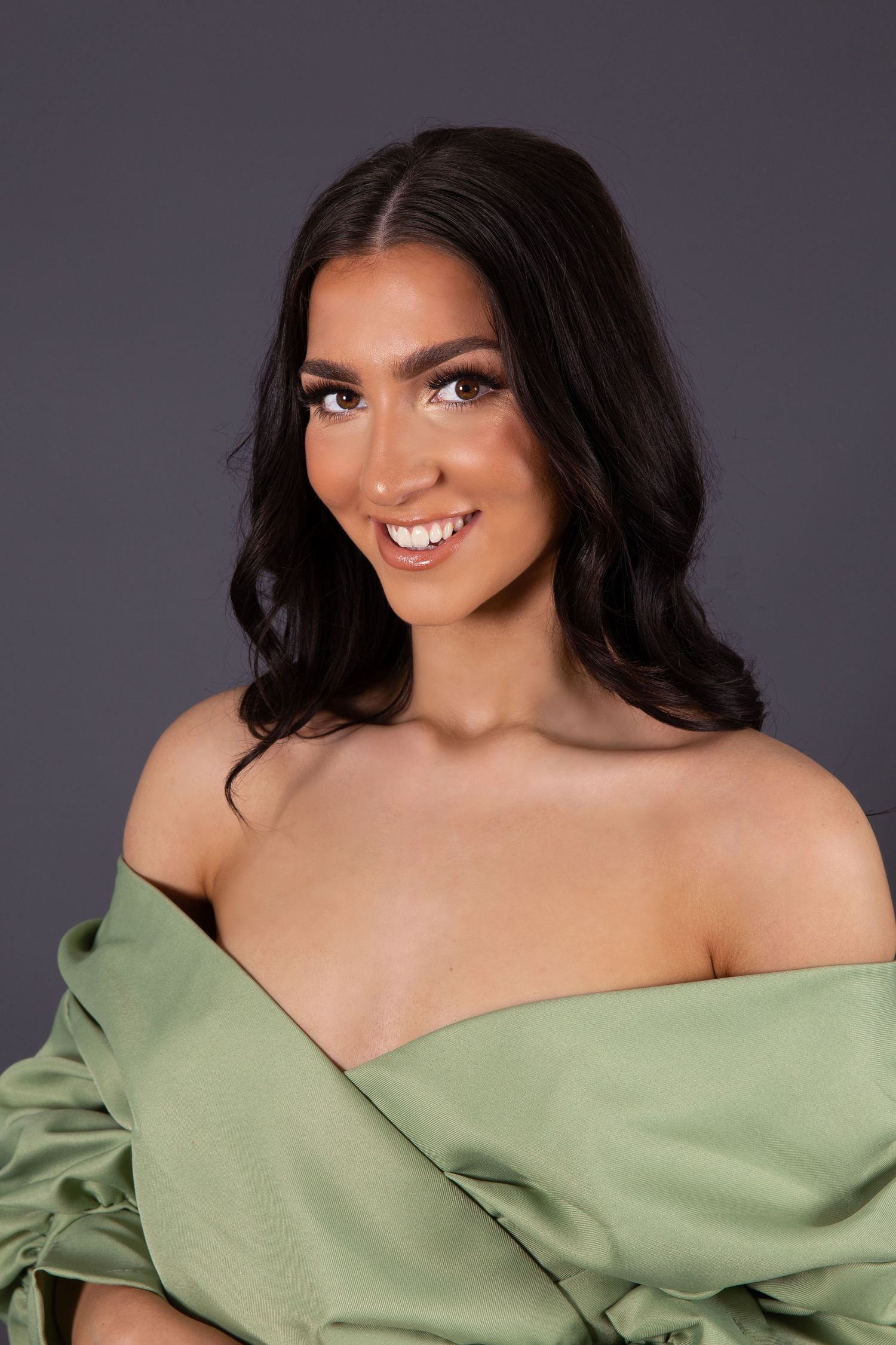 Portrait of a woman wearing an off-shoulder sage green top against a dark background, with dark wavy hair and a bright smile.