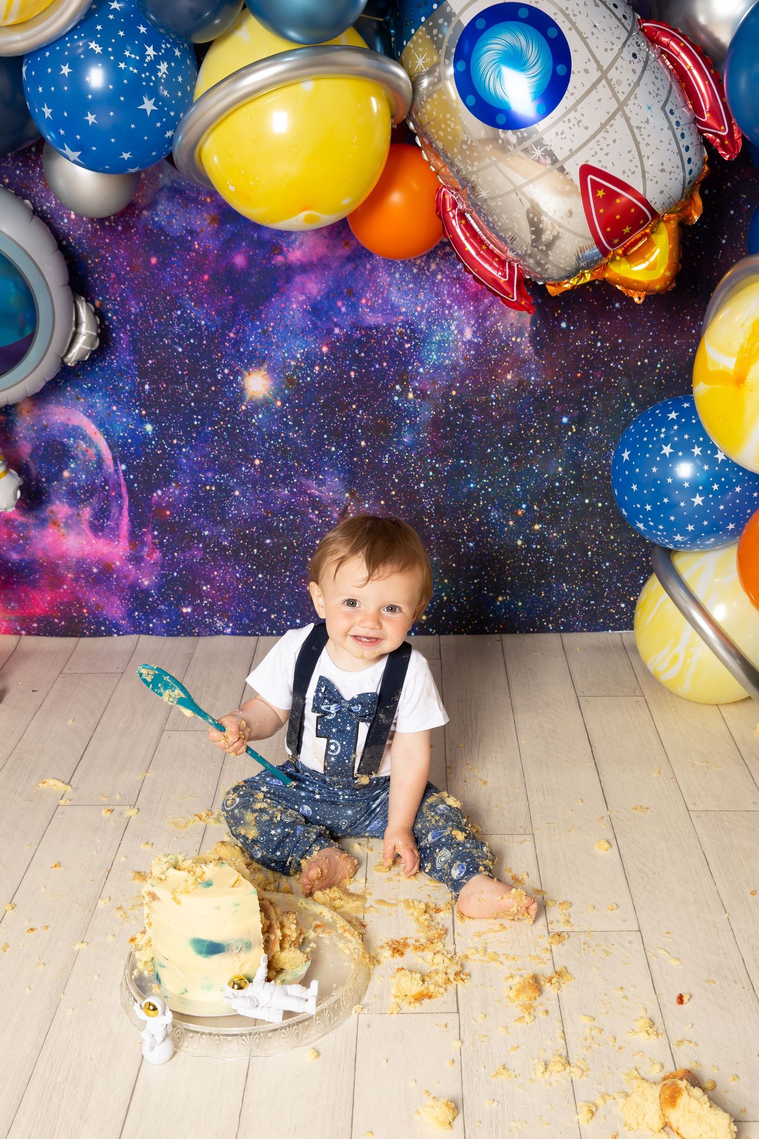 Fun space-themed cake smash photo setup with colorful planet balloons, galaxy backdrop and messy cake on wooden floor.