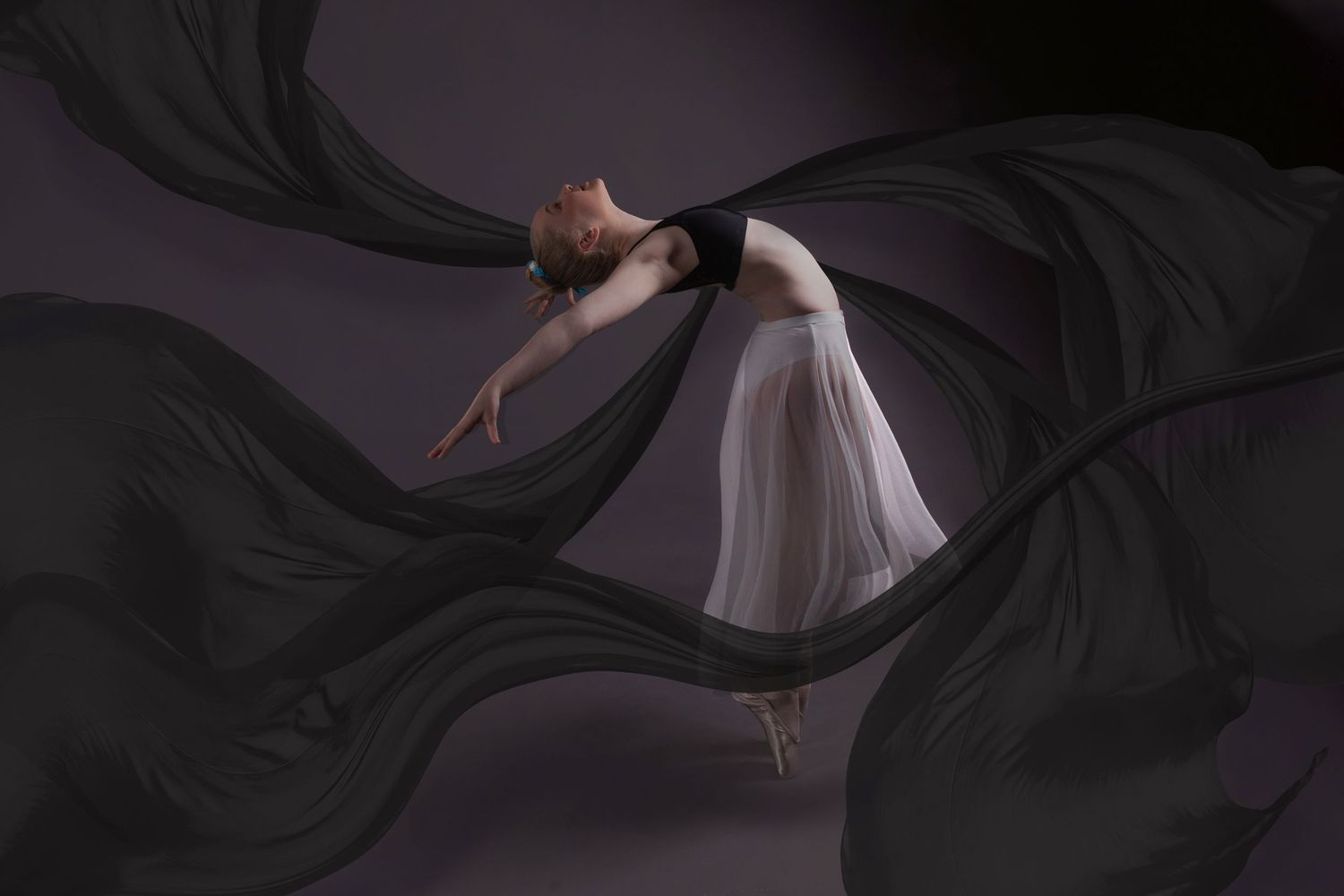 A graceful ballet dancer in a flowing white dress performs against a dramatic black background with billowing fabric.