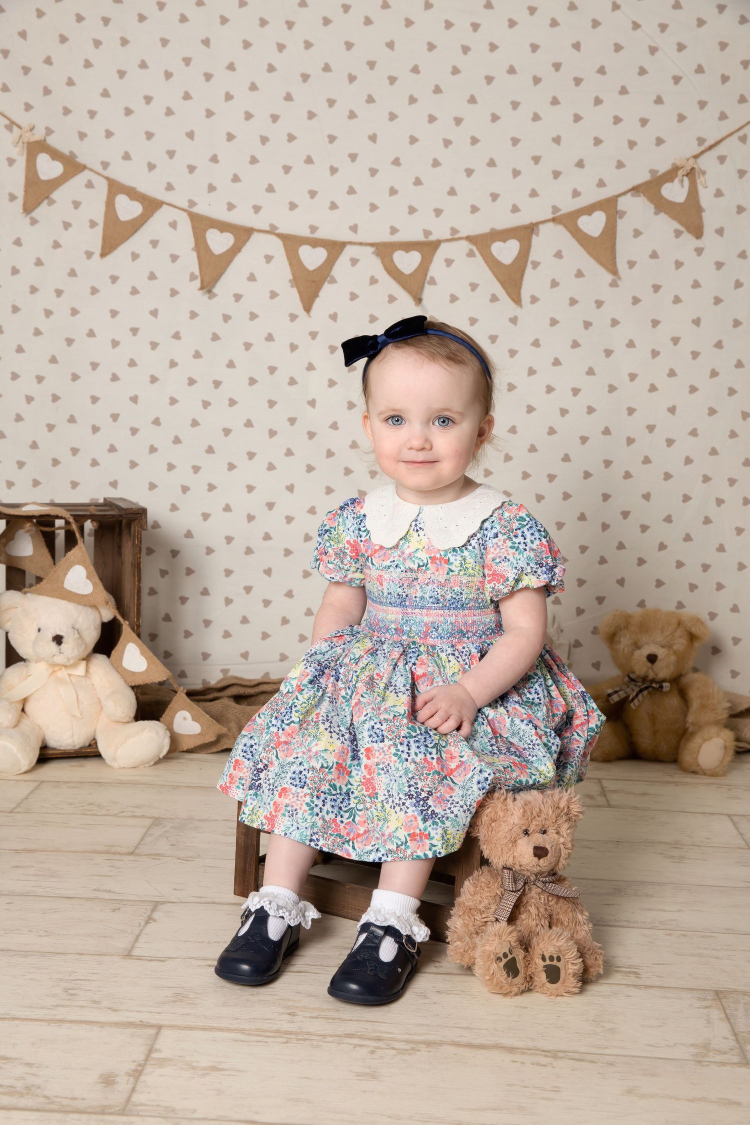 A small child in a floral dress sits on a wooden stool with teddy bears and decorative bunting in the background.