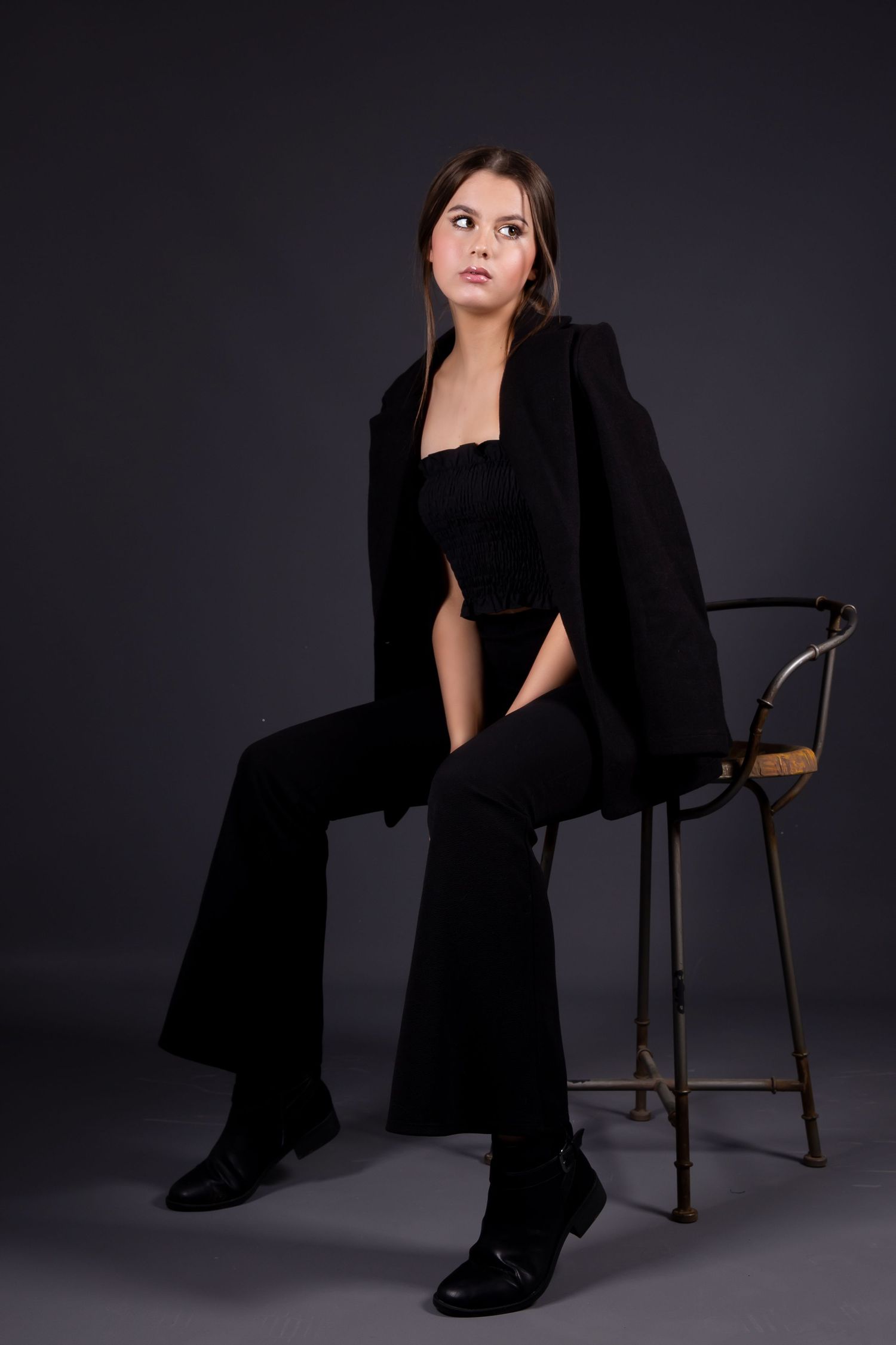 Model in all black attire including blazer and flare pants poses on chair against dark background in studio setting.