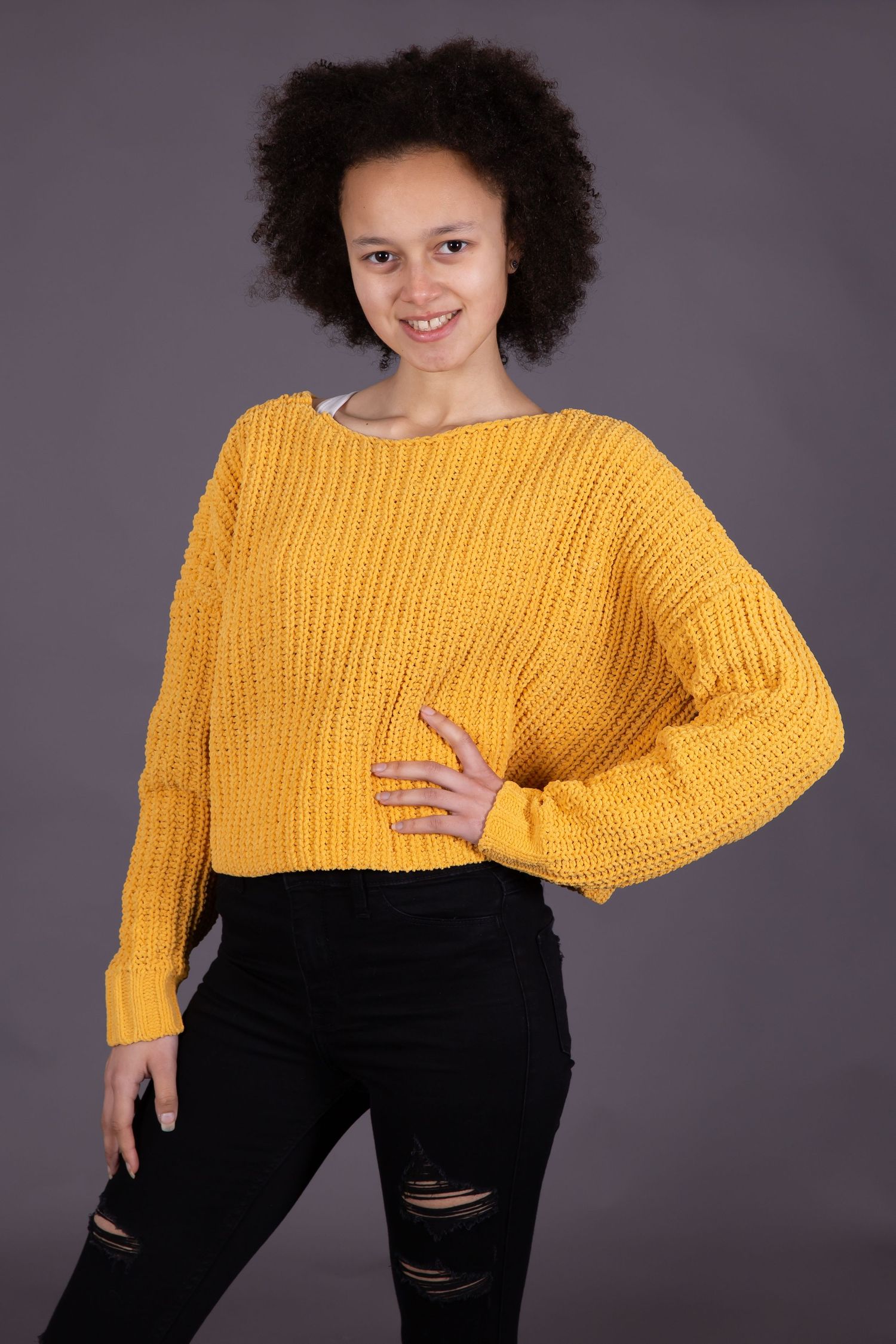 A person wearing a bright yellow chunky knit sweater and distressed black jeans poses against a gray background.