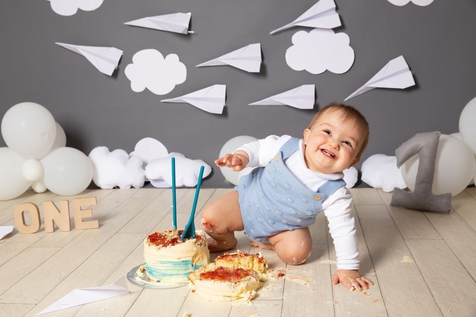 Baby in blue outfit smashes birthday cake during cake smash photo session with paper airplane and cloud decorations.