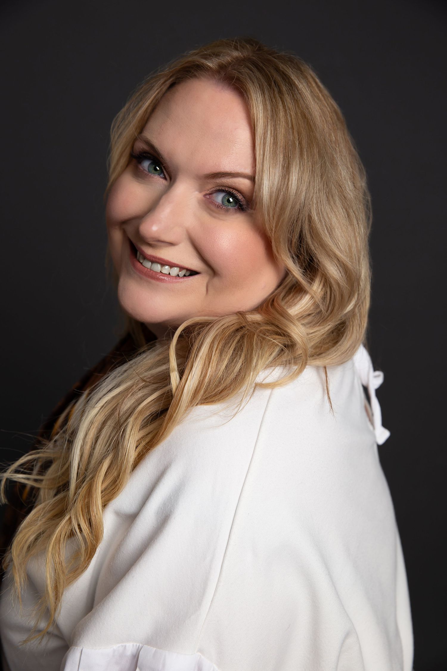 Professional studio portrait with blonde wavy hair wearing a white blazer against dark background, smiling warmly.