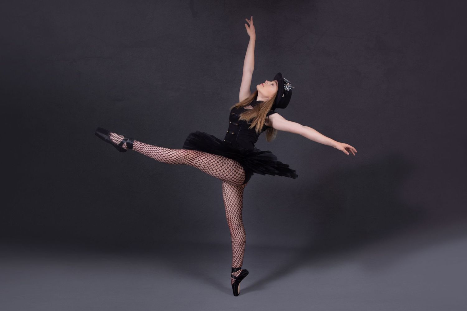 Ballerina in black tutu performs graceful arabesque pose against dark studio backdrop.