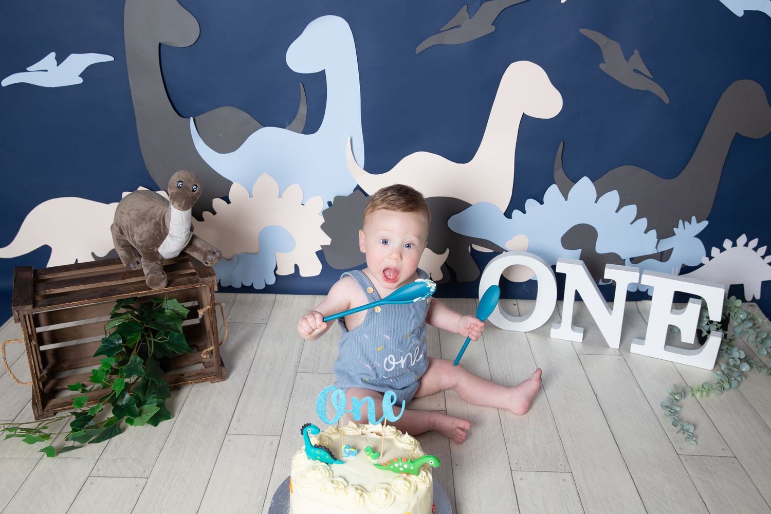 Dinosaur-themed first birthday party setup with wooden crate, decorative cutouts, and cake smash scene on light wooden floor.