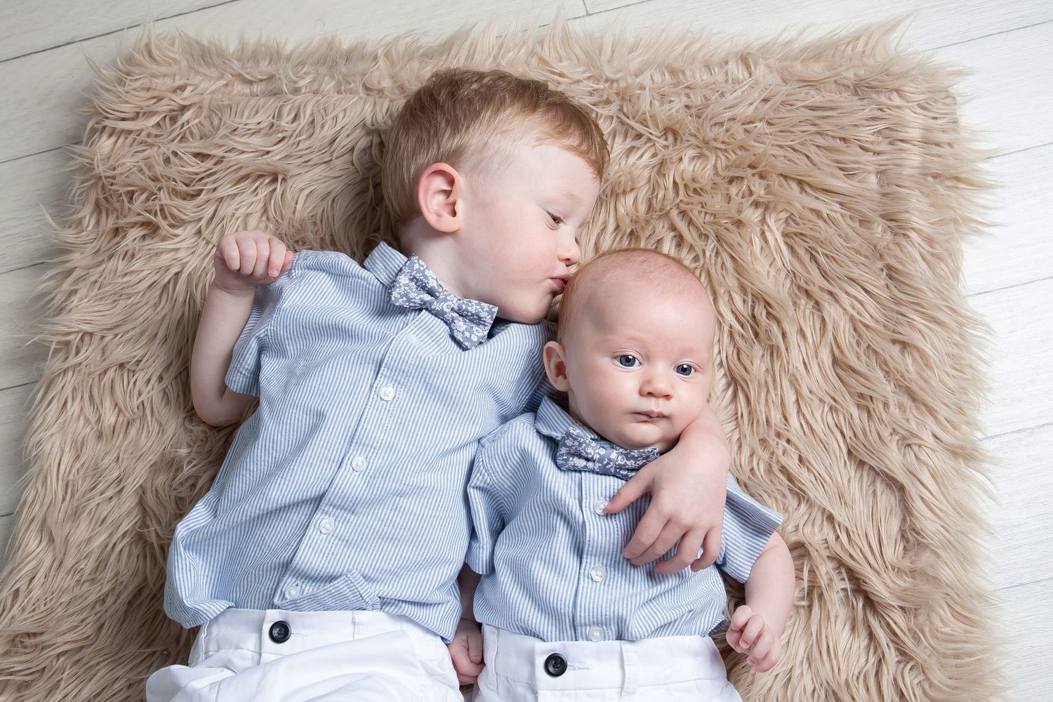 Two babies in matching light blue outfits lying together on a fluffy beige fur rug.