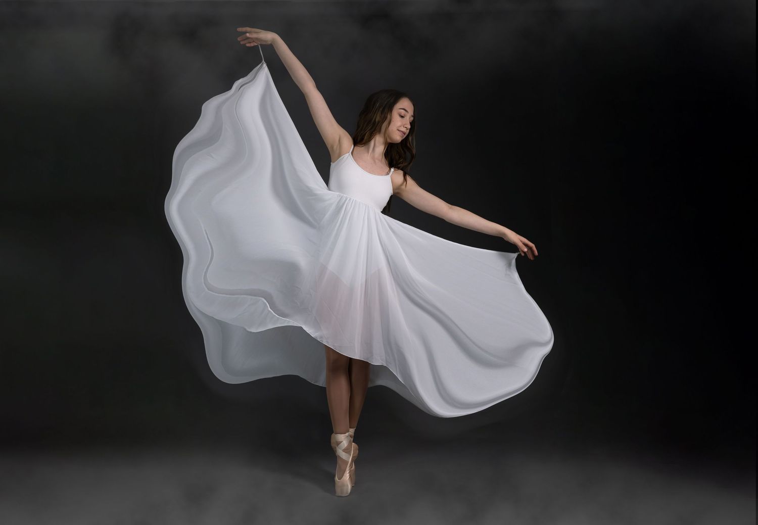 A dancer in a flowing white dress performs graceful ballet movements in a dramatic dark studio setting.