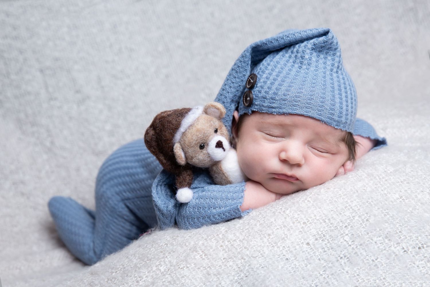 Sleeping newborn in blue knit outfit and hat cuddles small teddy bear on soft gray blanket.