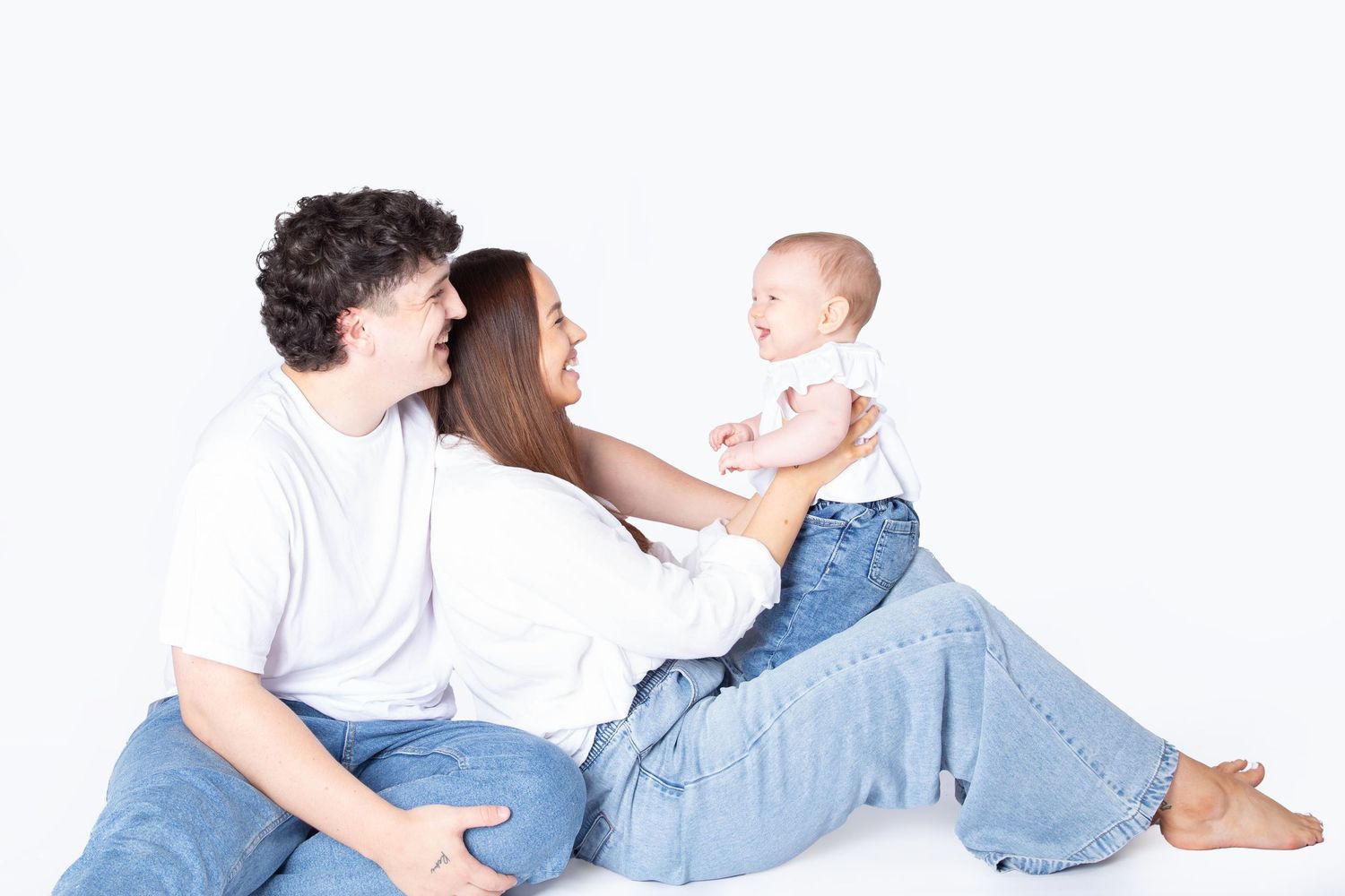 A couple in jeans and white shirts sits on white background with small child in denim playing between them.