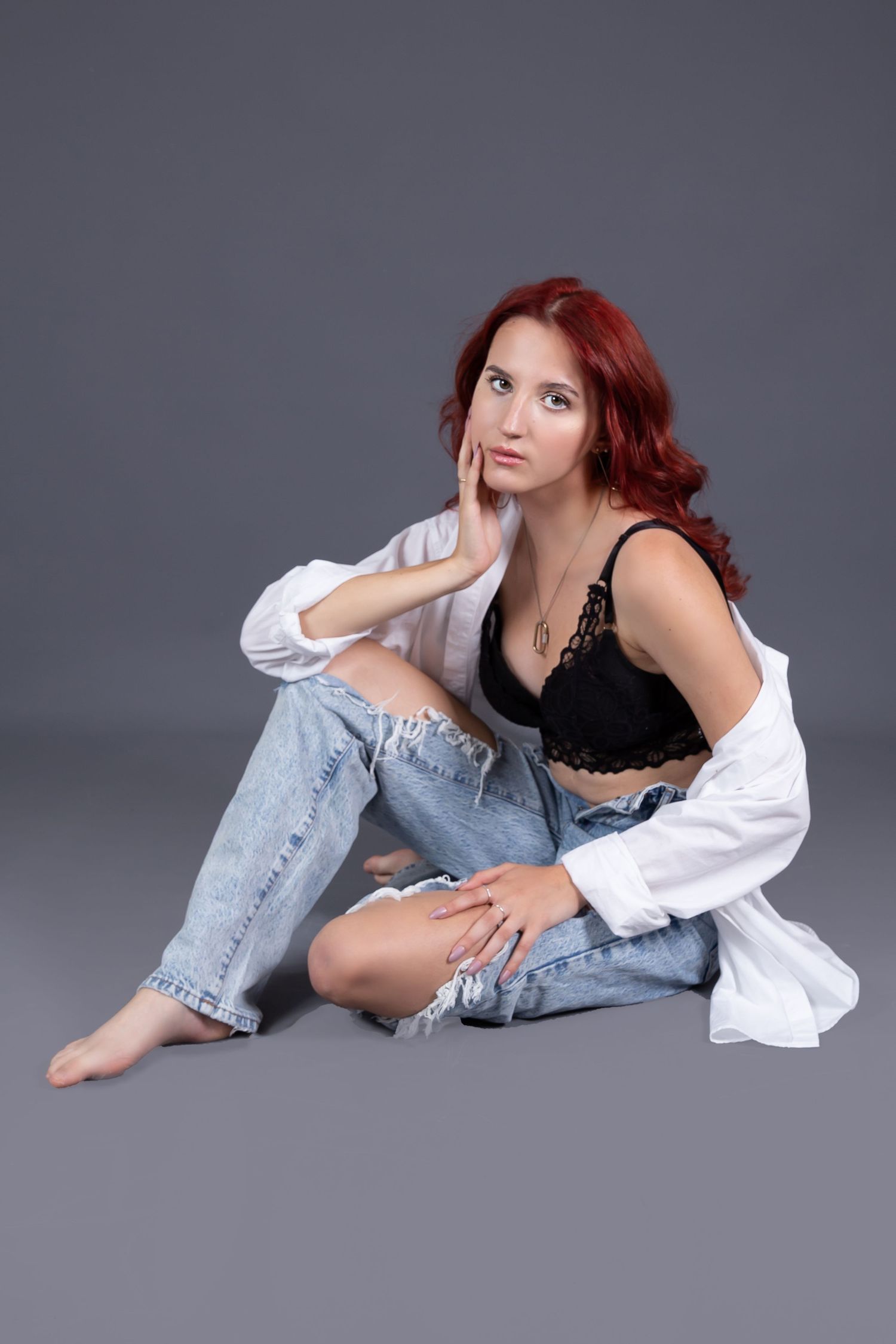 A person with red hair wearing a white shirt and jeans lounges on a gray studio floor in artistic poses.
