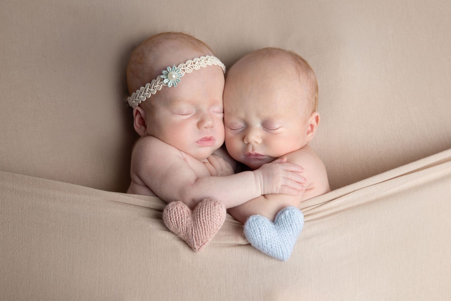 Two sleeping newborns cuddling together on beige fabric with pink and blue heart decorations.