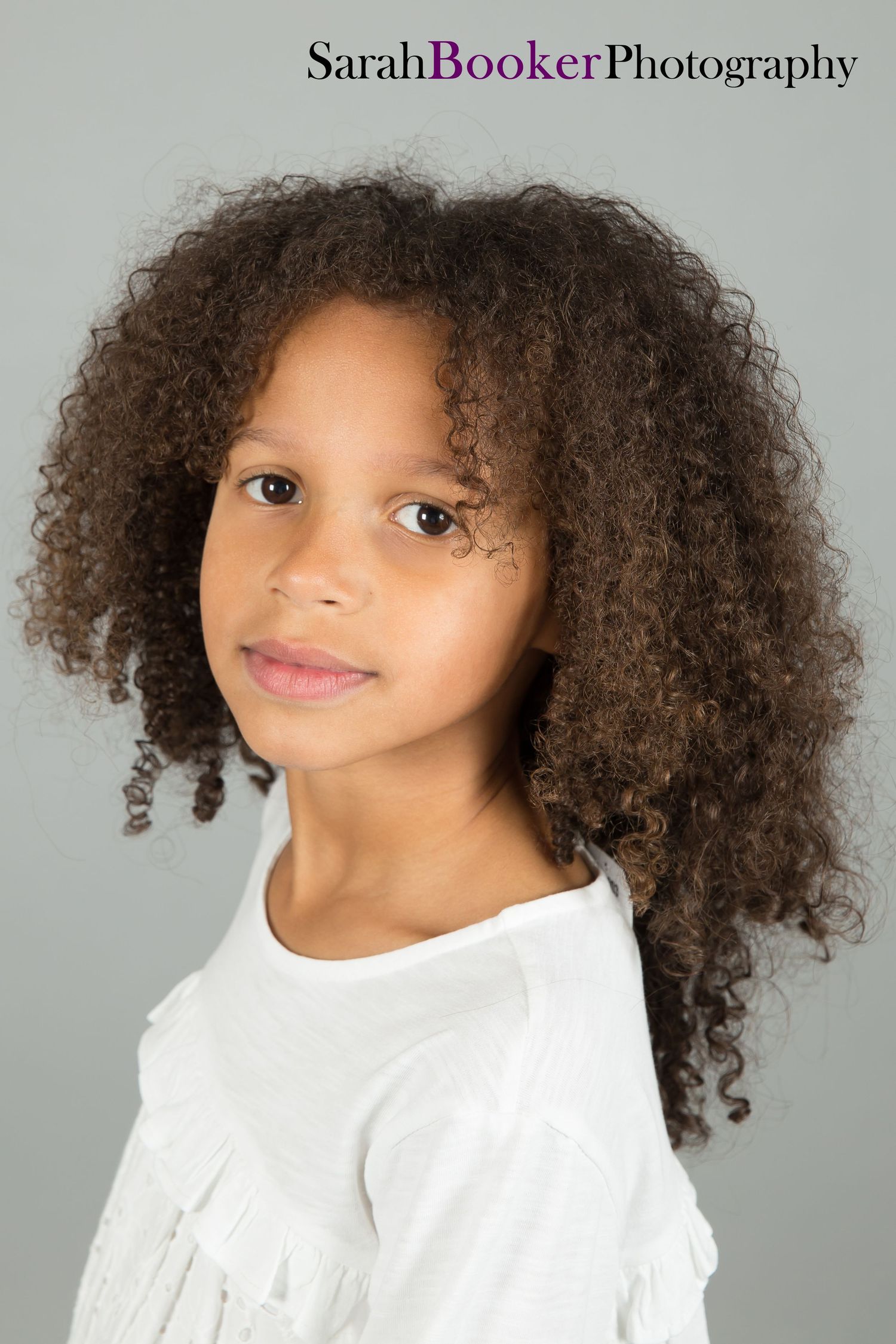 Professional studio portrait of a young person with natural curly hair wearing a white top against a gray background.