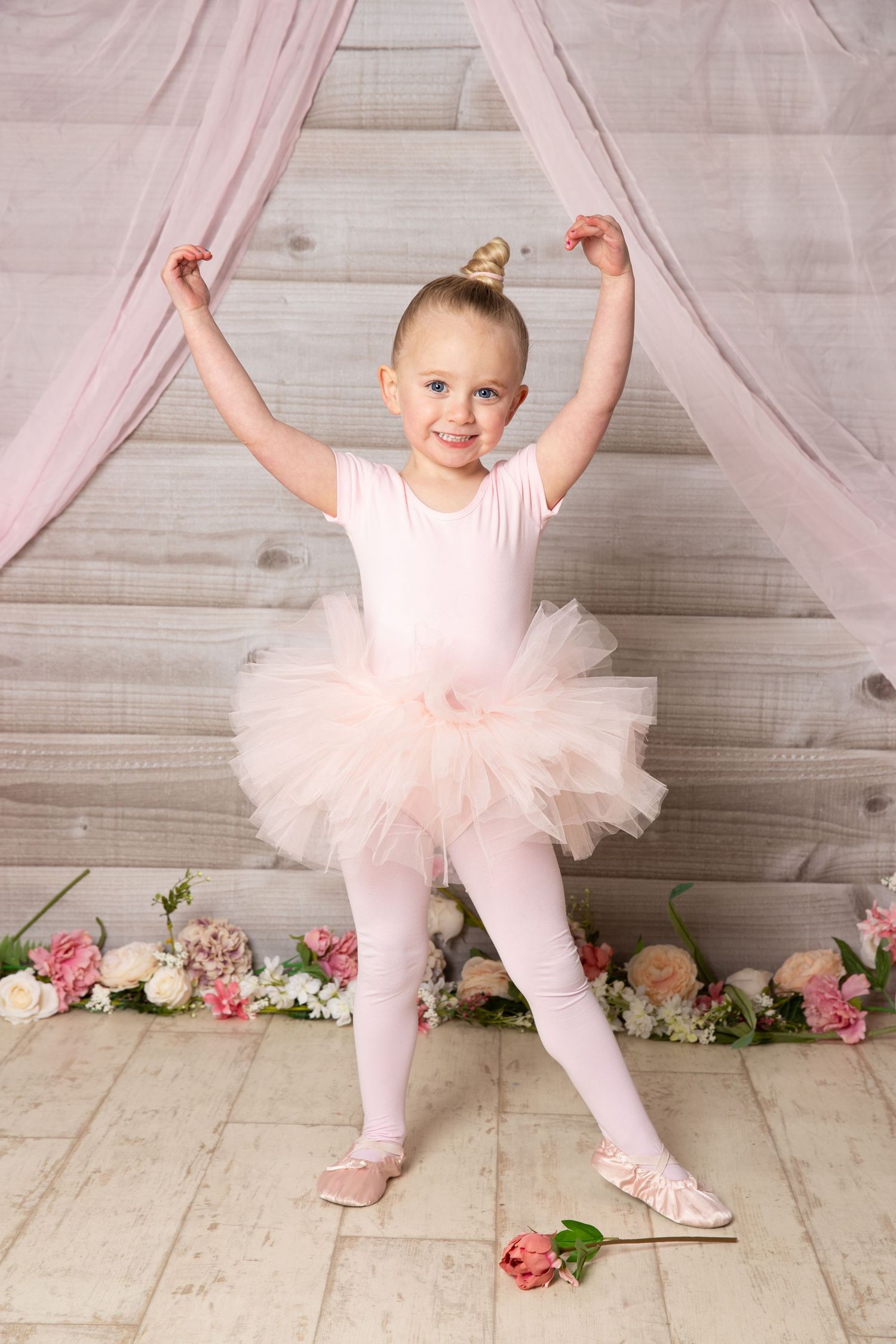 A young ballet dancer in pink tutu and tights poses joyfully against a rustic backdrop with scattered pink roses on the floor.