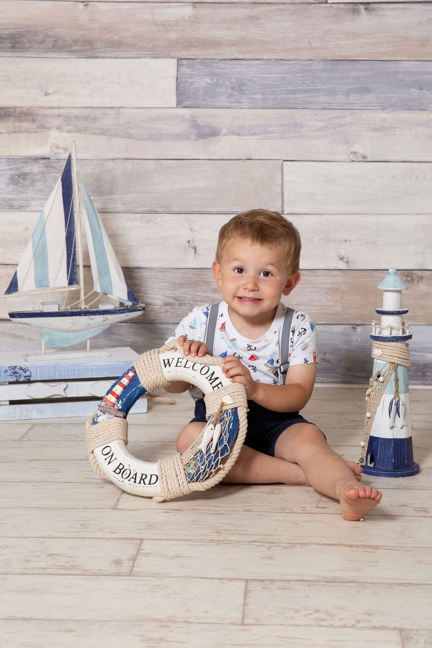 Nautical themed photo setup with a decorative life preserver ring, toy sailboat and lighthouse on wooden plank background.