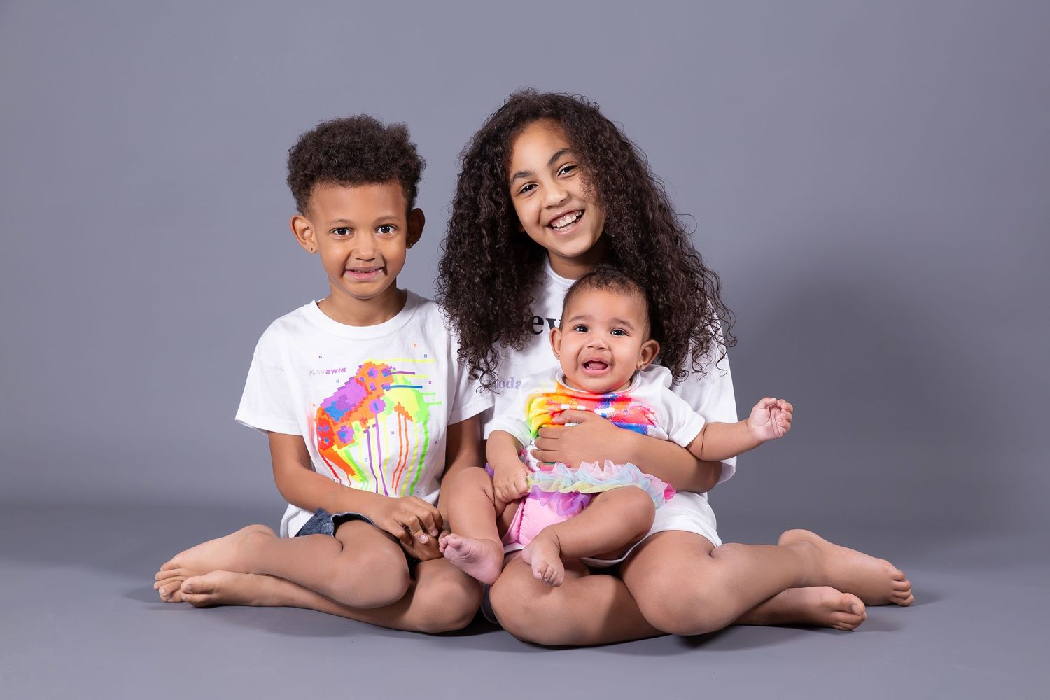 Two small children in white outfits sit together on a gray backdrop playing with colorful toy balls.