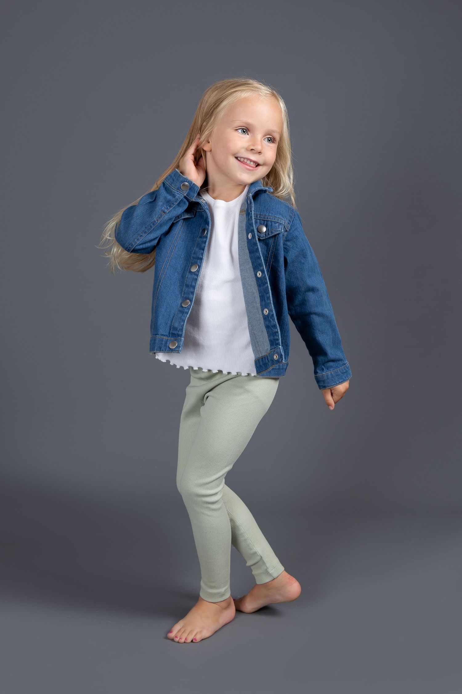 Dancing child wearing denim jacket, white top and mint green pants poses barefoot against gray background.