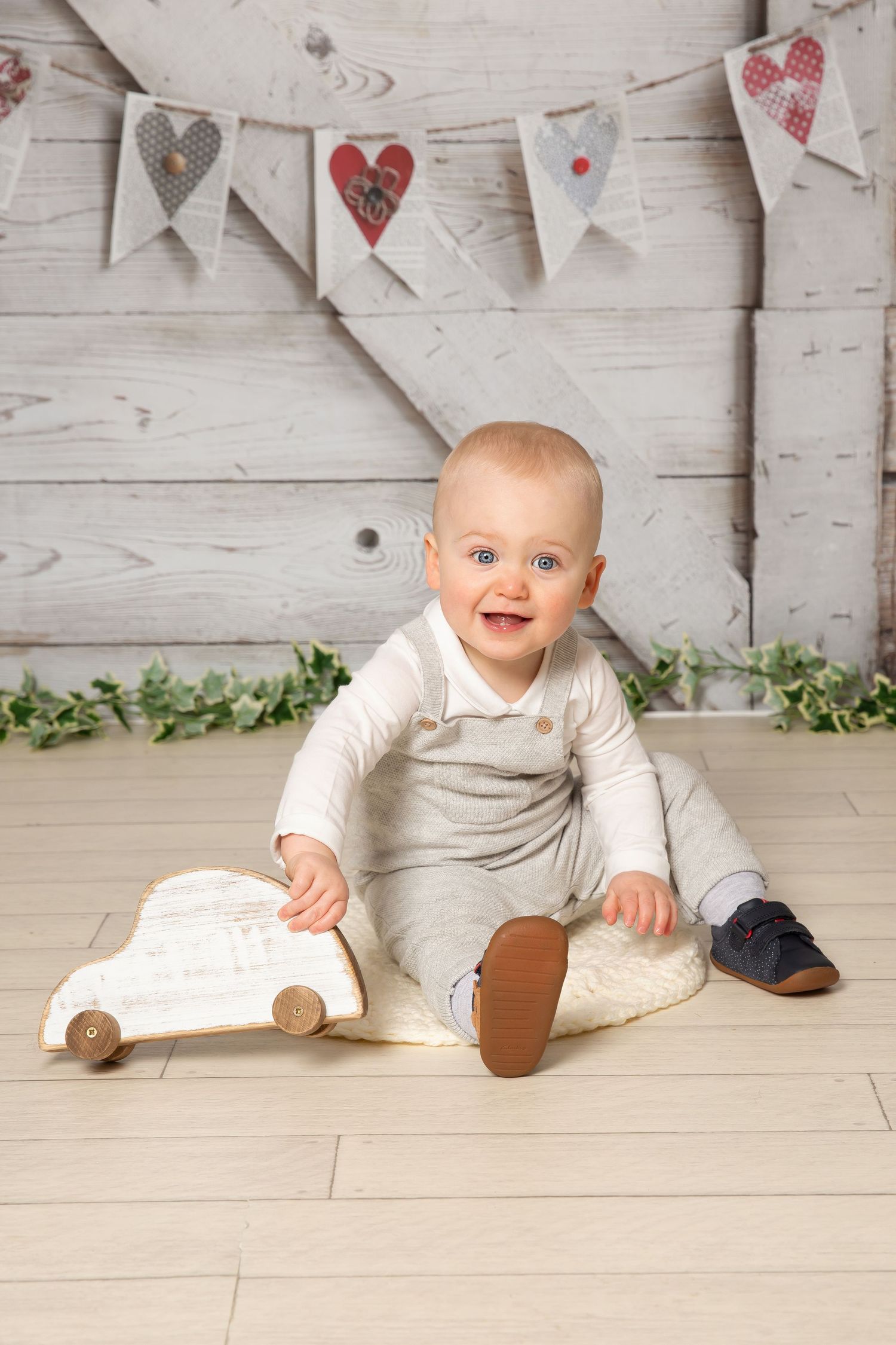 Small baby wearing white outfit sits on wooden floor playing with toy car under heart banner decorations.
