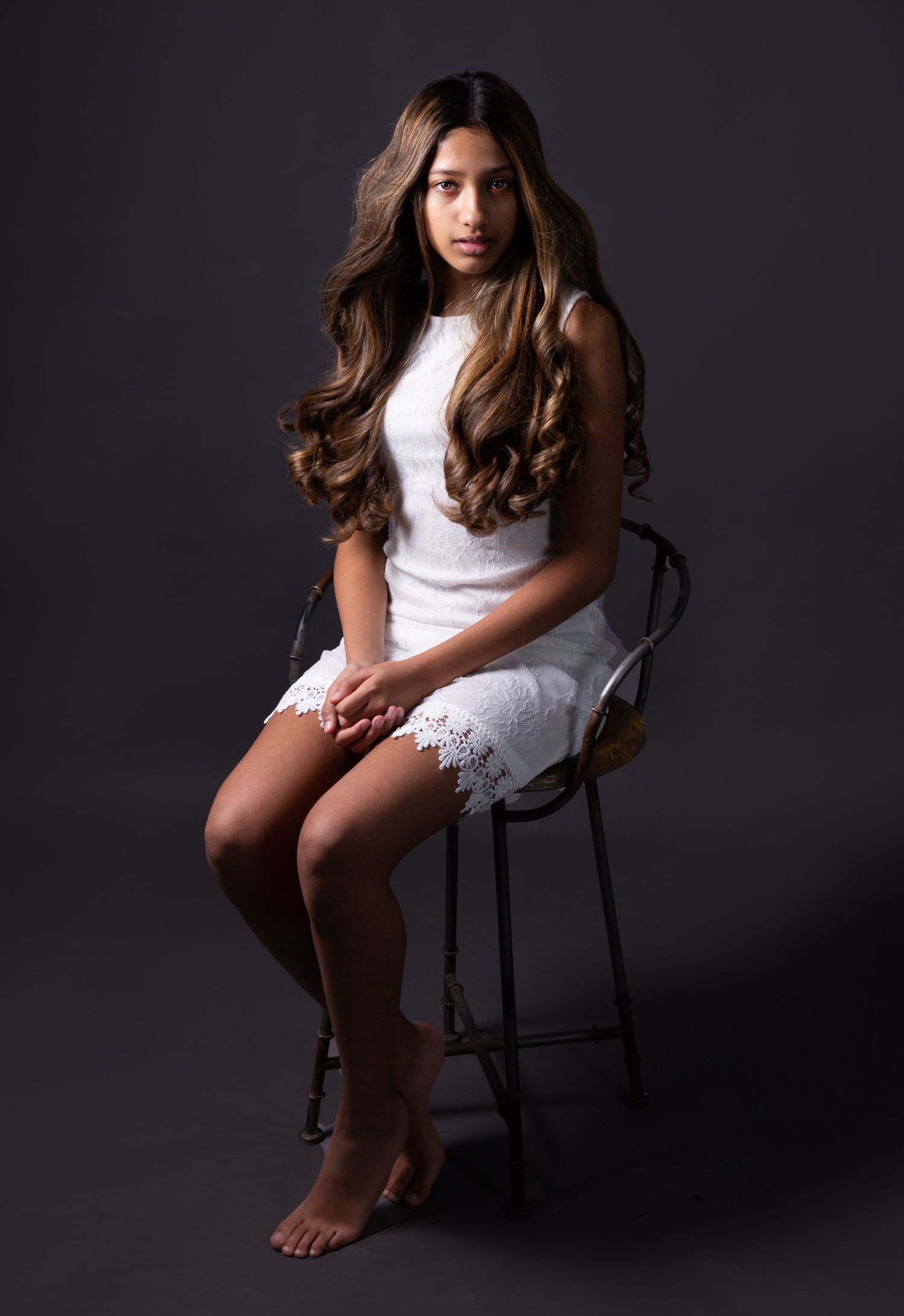 Portrait of someone sitting on a dark stool wearing a white lace dress against a black background in moody lighting.