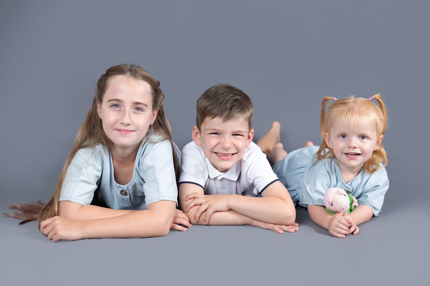 Three siblings lying comfortably on studio floor with gray backdrop during family photo shoot.