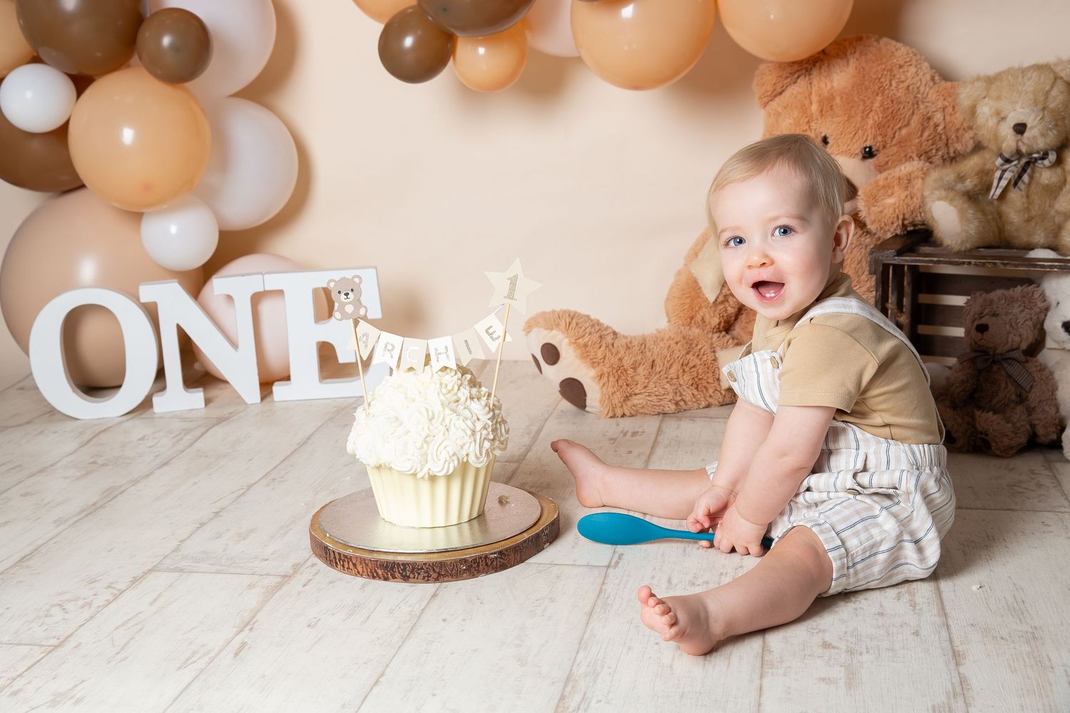 Baby in white outfit sits near neutral-colored balloon arch with cake and decorations for first birthday celebration.
