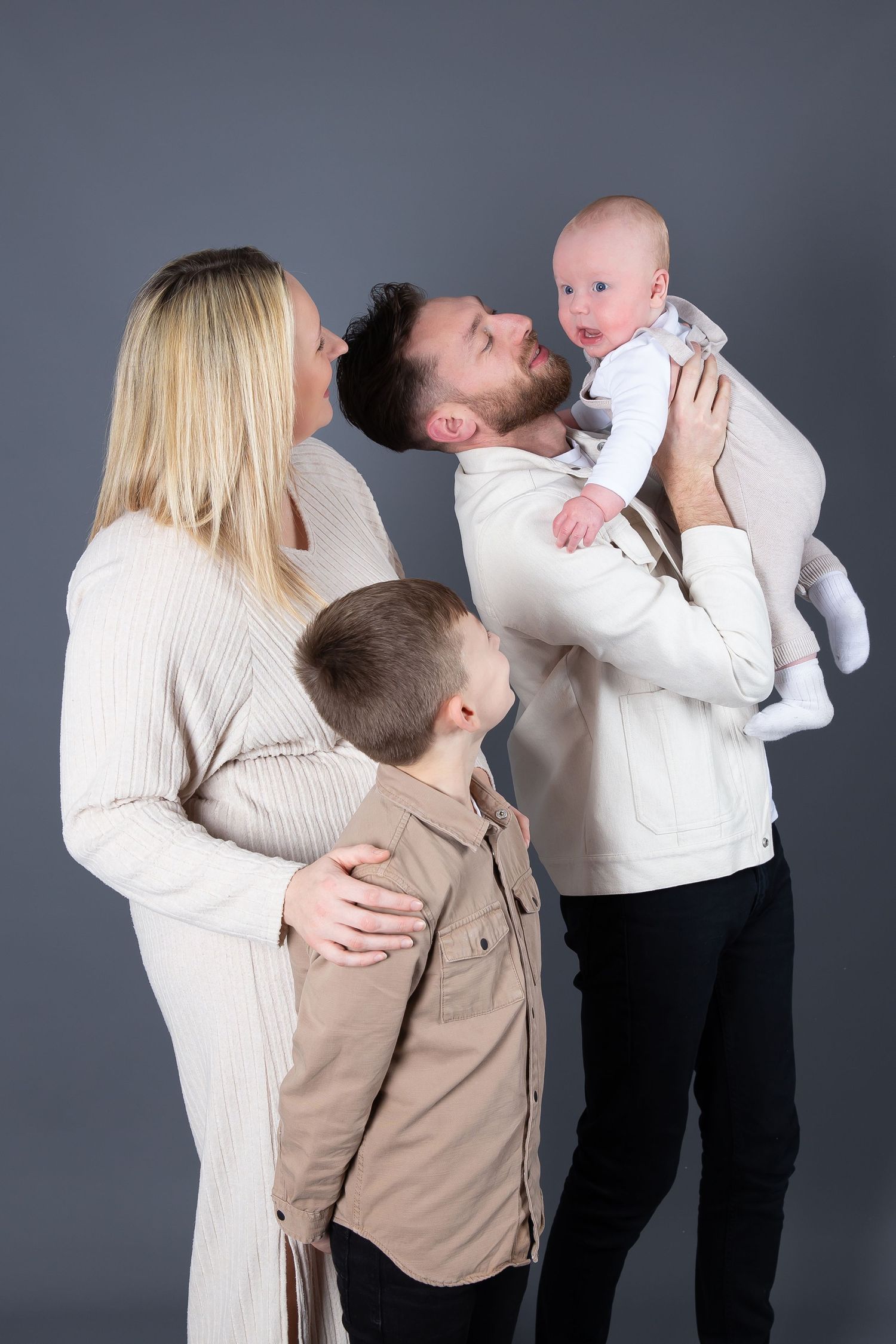 A family in white and beige outfits poses together against a gray backdrop for a professional studio portrait.