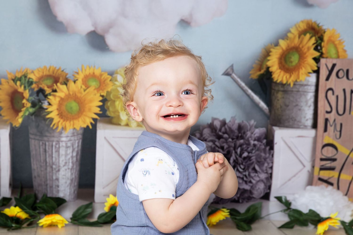 Cheerful toddler sits among decorative sunflowers in galvanized buckets against a gray backdrop.